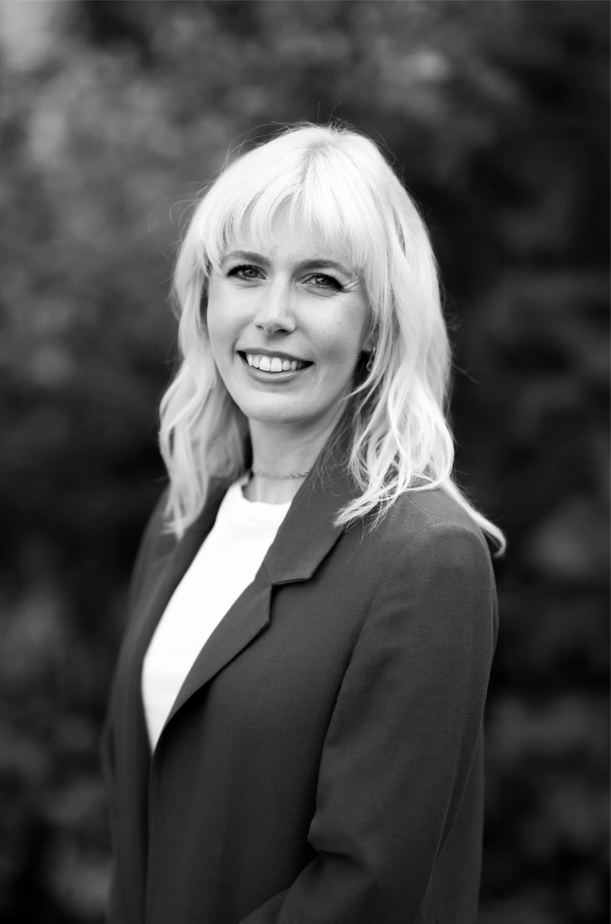Black and white portrait of a smiling woman with shoulder-length blonde hair, wearing a blazer and a white top, outdoors with blurred foliage in the background.