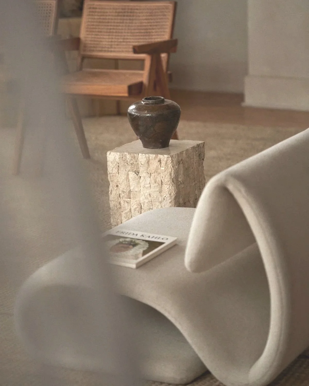 A small dark pottery vase on a textured stone pedestal in a room with wooden furniture and a magazine on a white fabric surface.