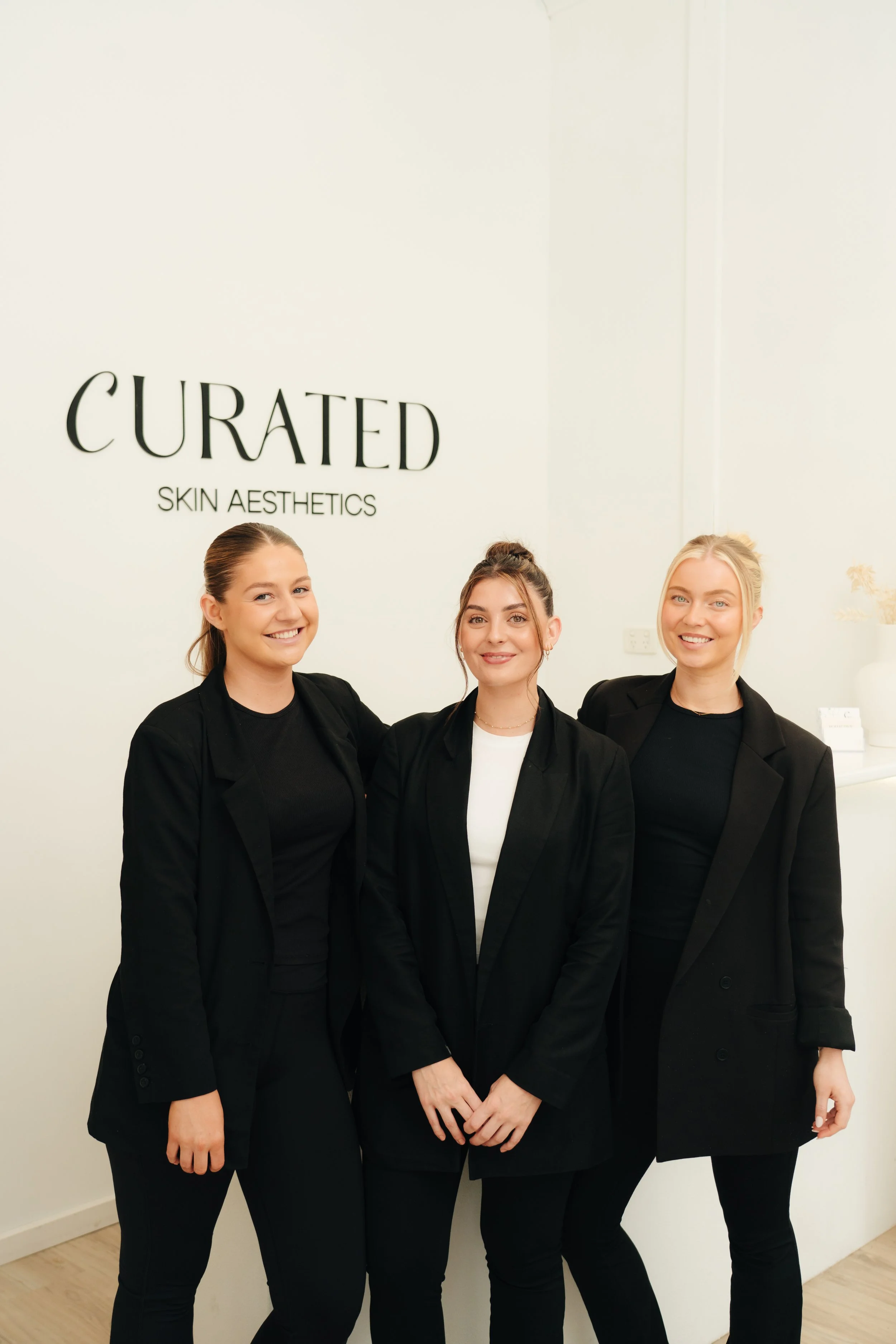 Three women in black blazers smiling in front of a white wall with 'Curated Skin Aesthetics' sign.