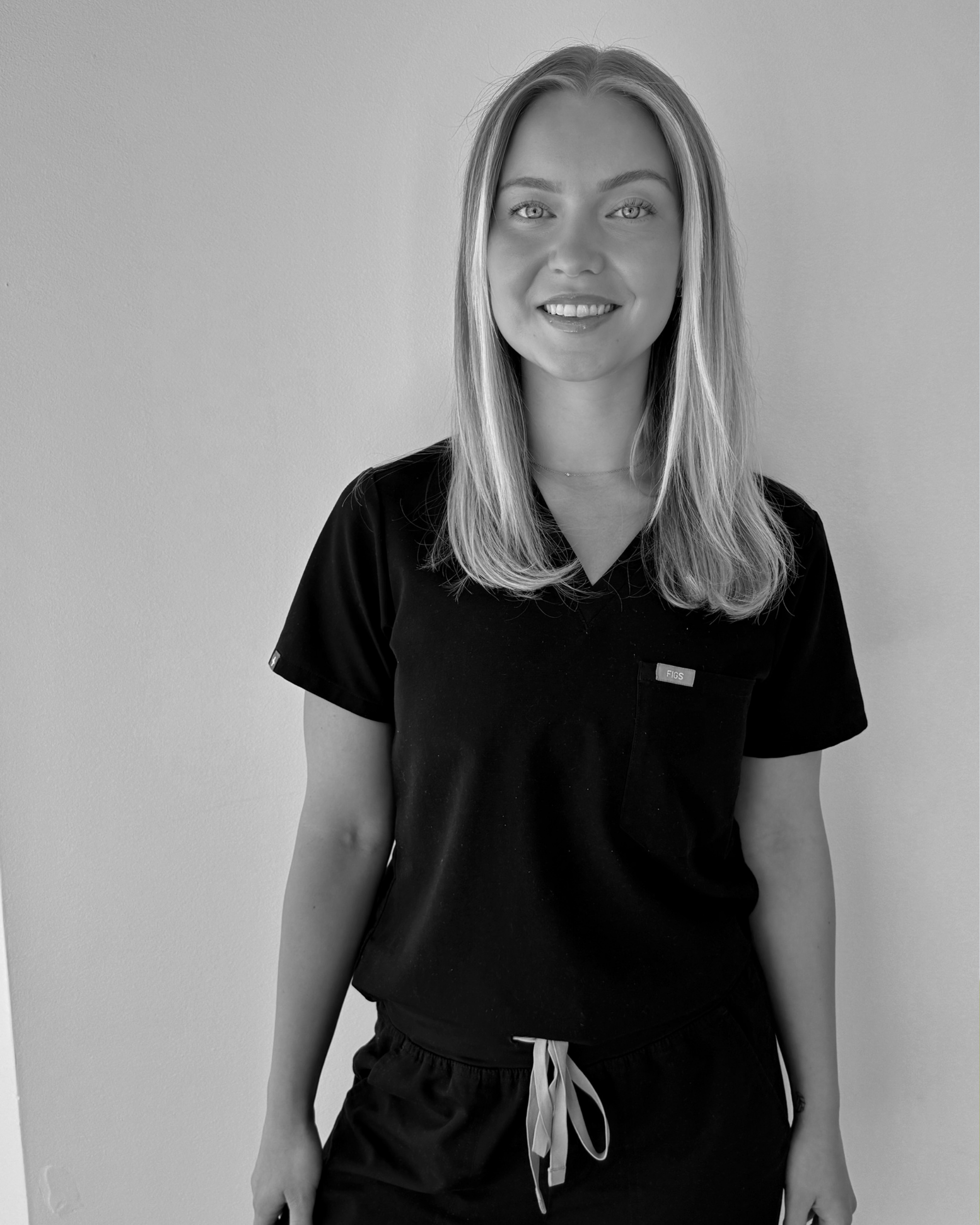 Black and white photograph of a young woman with shoulder-length hair, smiling and wearing a dark scrubs top and drawstring shorts, standing against a plain wall.