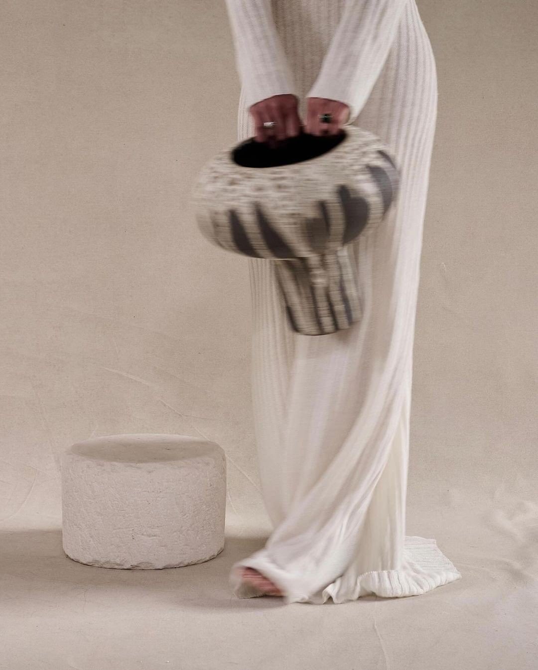 Person in a long, white, striped dress placing a striped, abstract-shaped object on the ground next to a small, round, white textured platform.