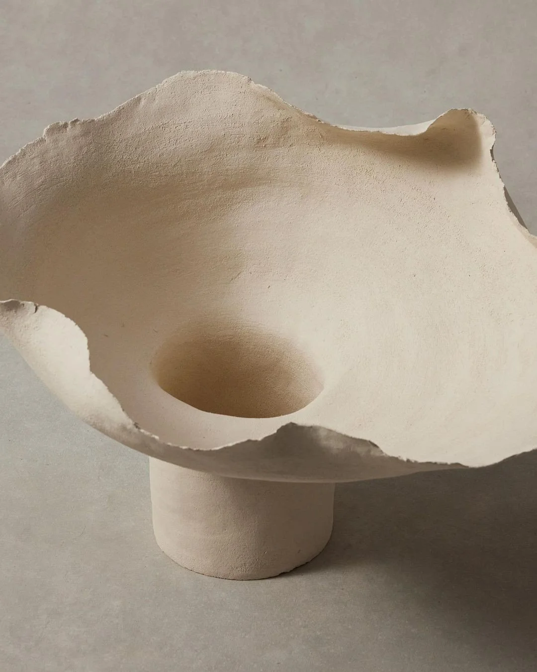A white ceramic or clay bowl with an irregular shape, resembling a flower or shell, mounted on a pedestal base.