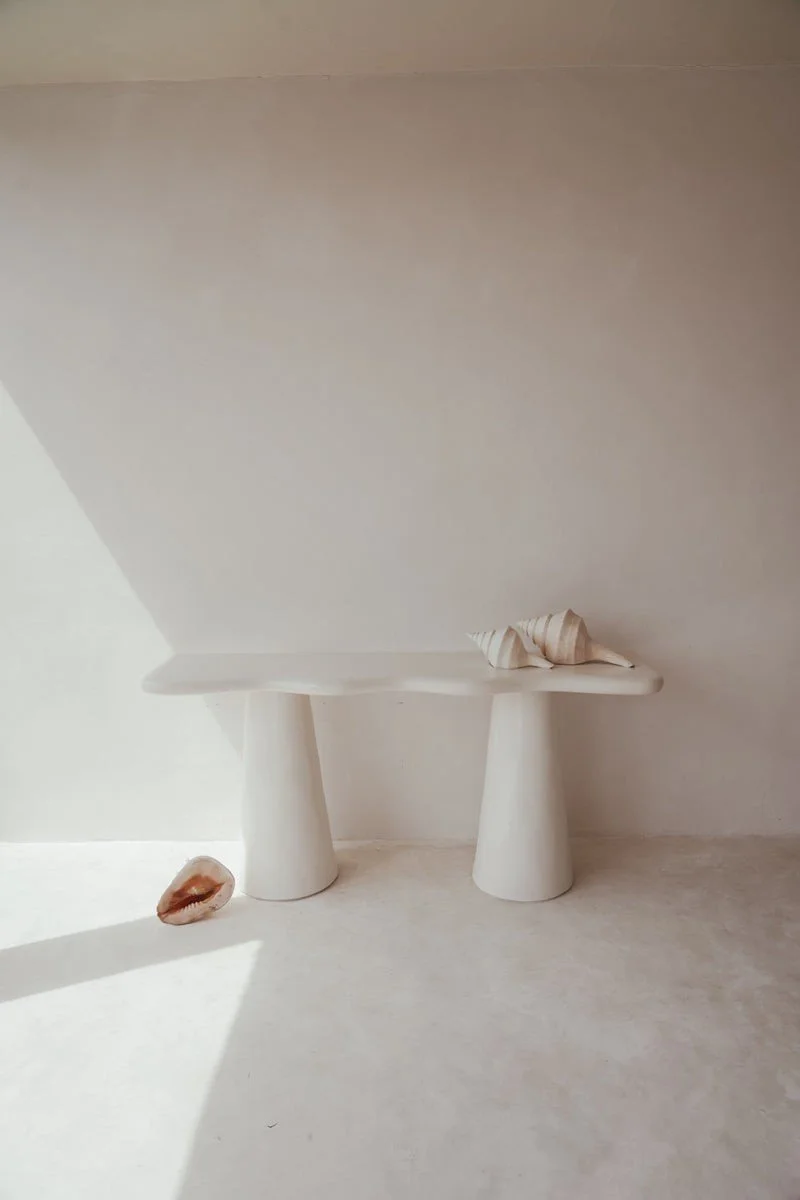 Minimalist scene with a white concrete bench supported by two conical legs, two shells on the bench, one shell on the floor, and plain white walls and floor.