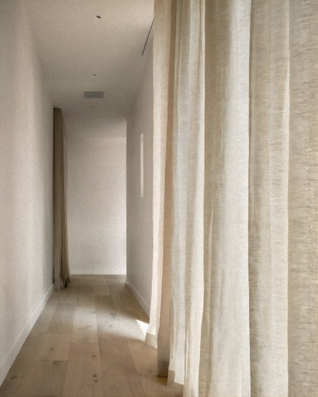 A minimalist hallway with light-colored walls, wooden flooring, and beige curtains hanging along the right side.