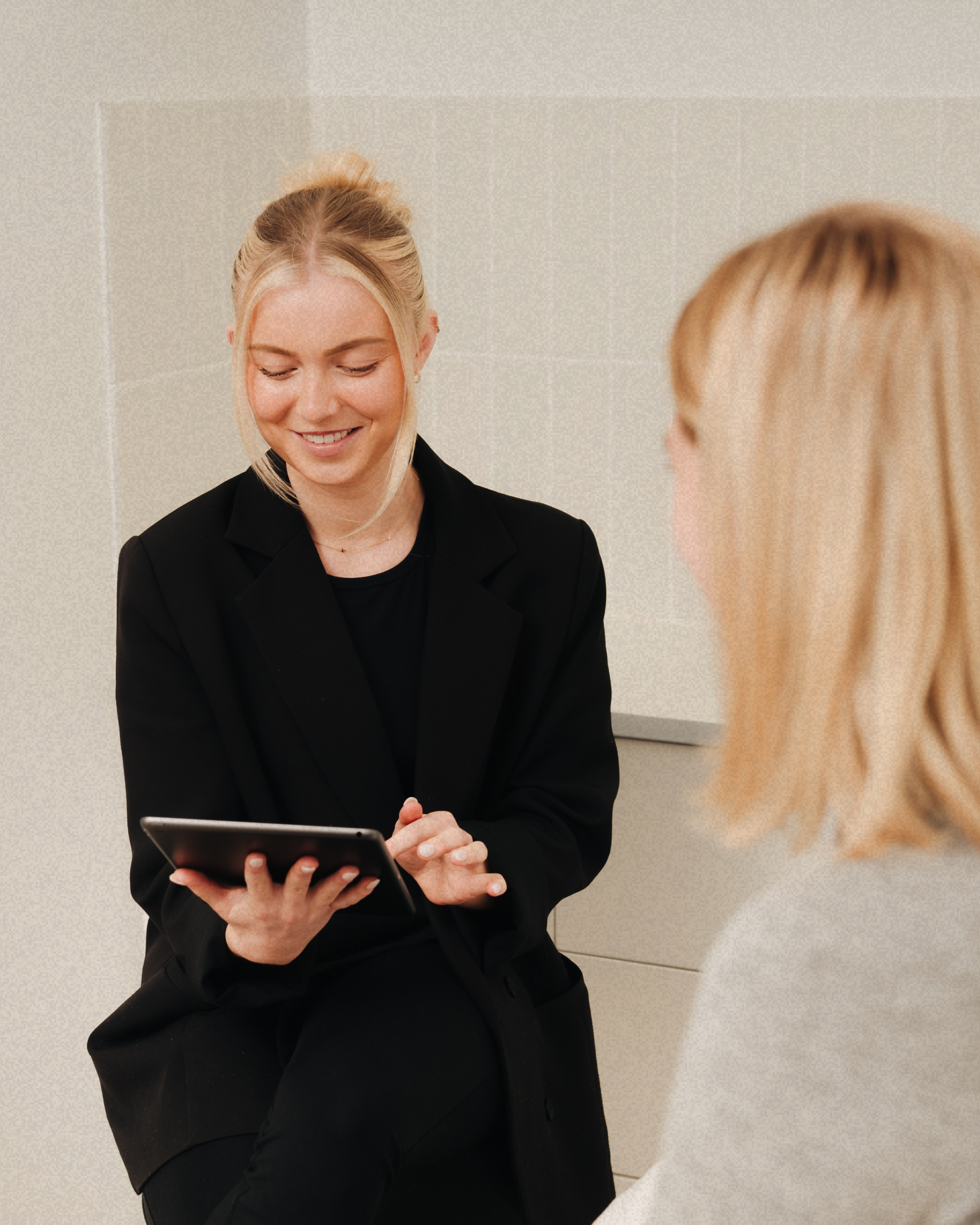 Two women having a conversation in an office, one is showing something on a tablet to the other, both smiling.
