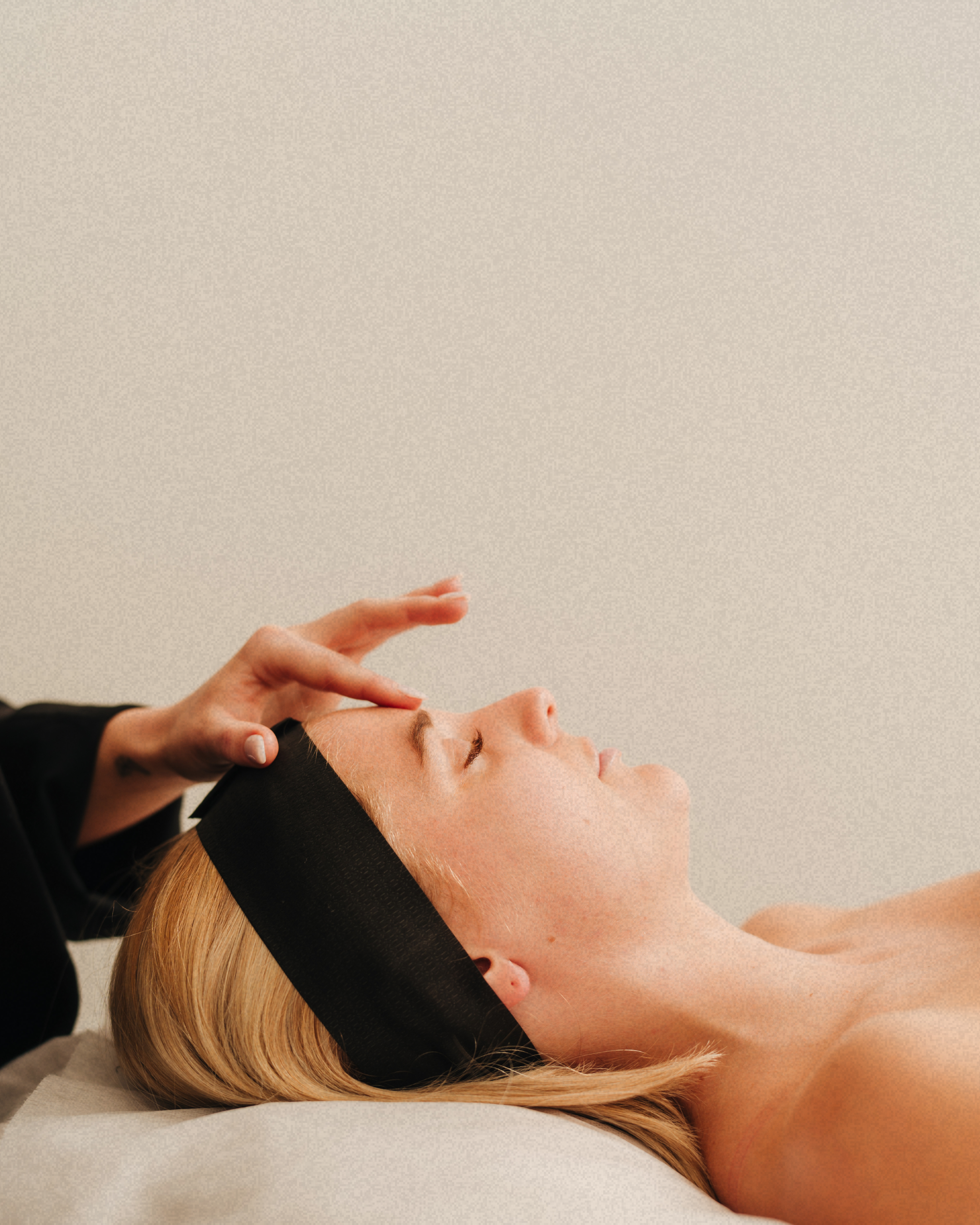 A woman lying down with eyes closed receiving a facial treatment from a professional, who is wearing a black headband and black clothing, applying a product to her forehead.