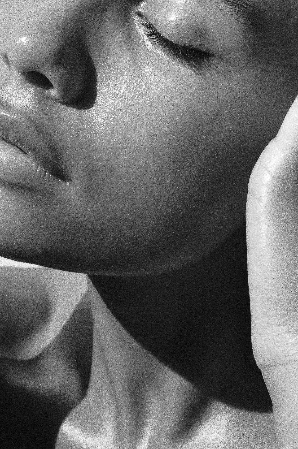 Close-up black and white photograph of a person's face, showing closed eyes, nose, and lips, with visible skin texture and a hand resting on the cheek.