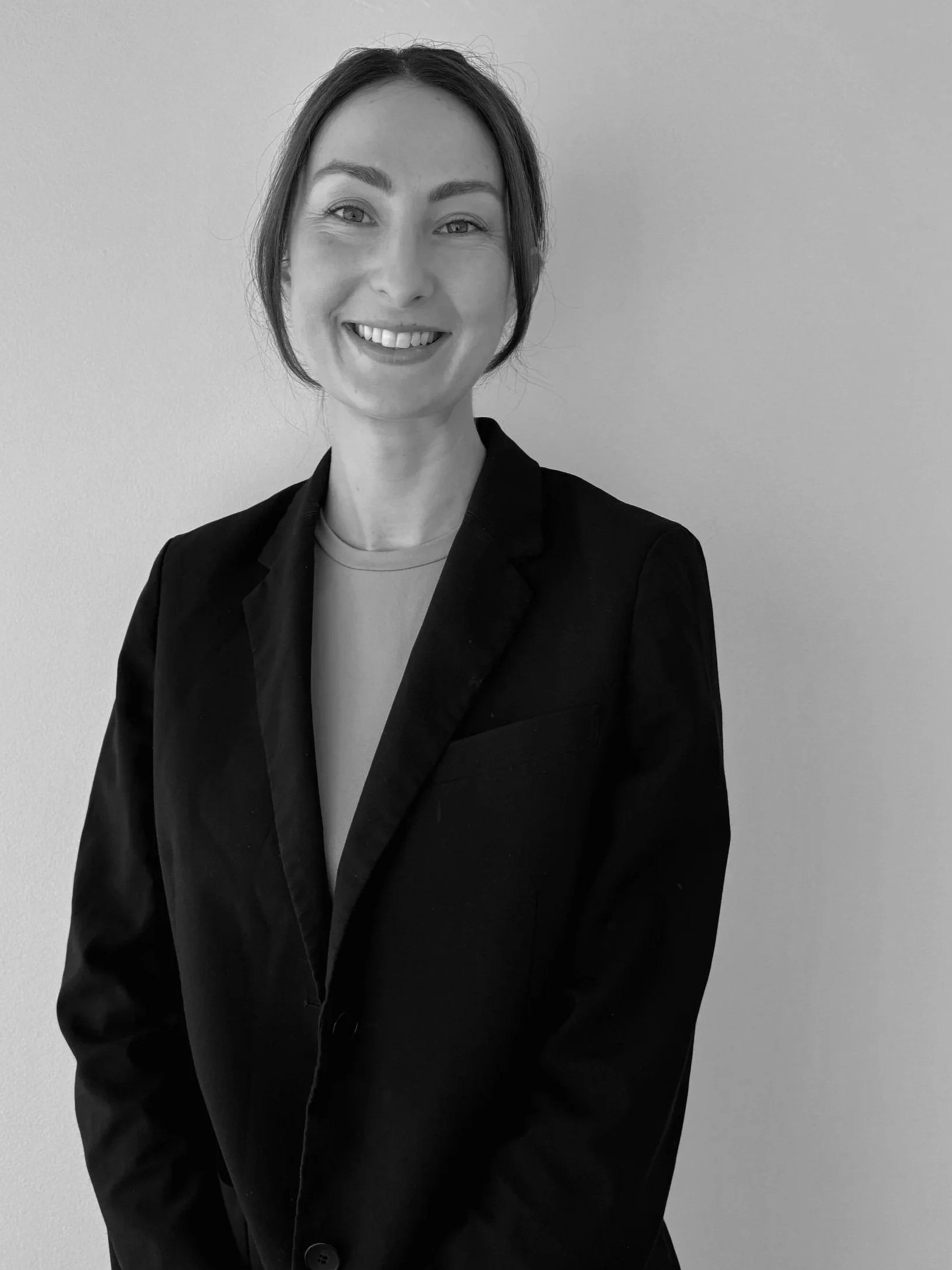 Black and white portrait of a woman smiling, wearing a blazer and a light-colored top, standing against a plain light-colored wall.