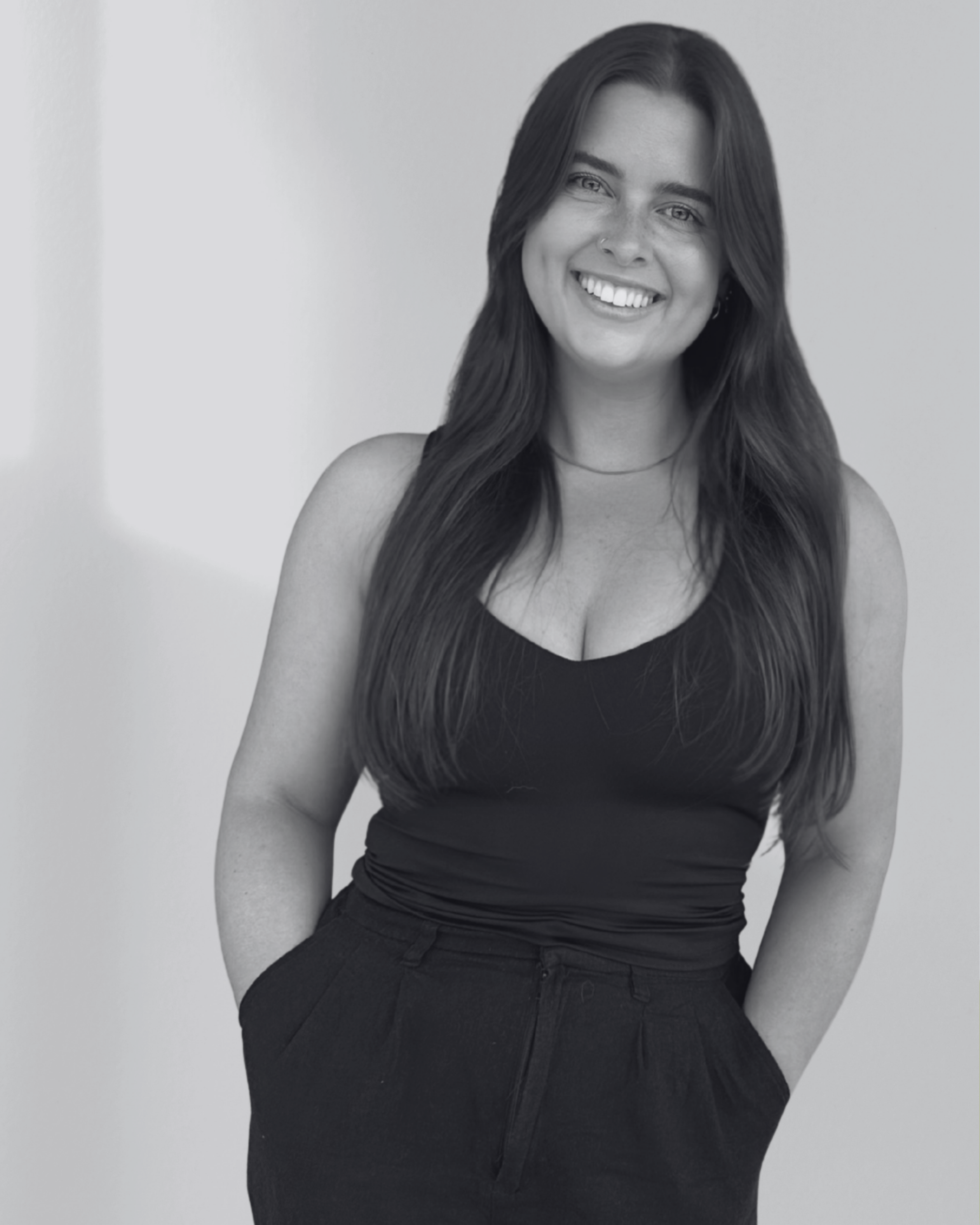 A young woman with long dark hair smiling, wearing a sleeveless black top and high-waisted black pants, standing against a plain light background.