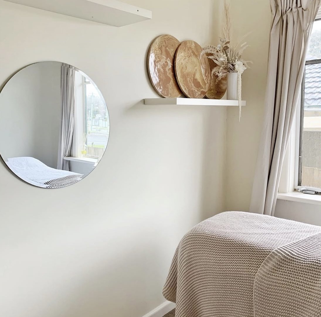 A cozy bedroom corner featuring a round mirror mounted on a white wall, a white floating shelf holding decorative brown trays and a white vase with dried flowers, a window with cream curtains letting in natural light, and a bed with textured beige bedding.