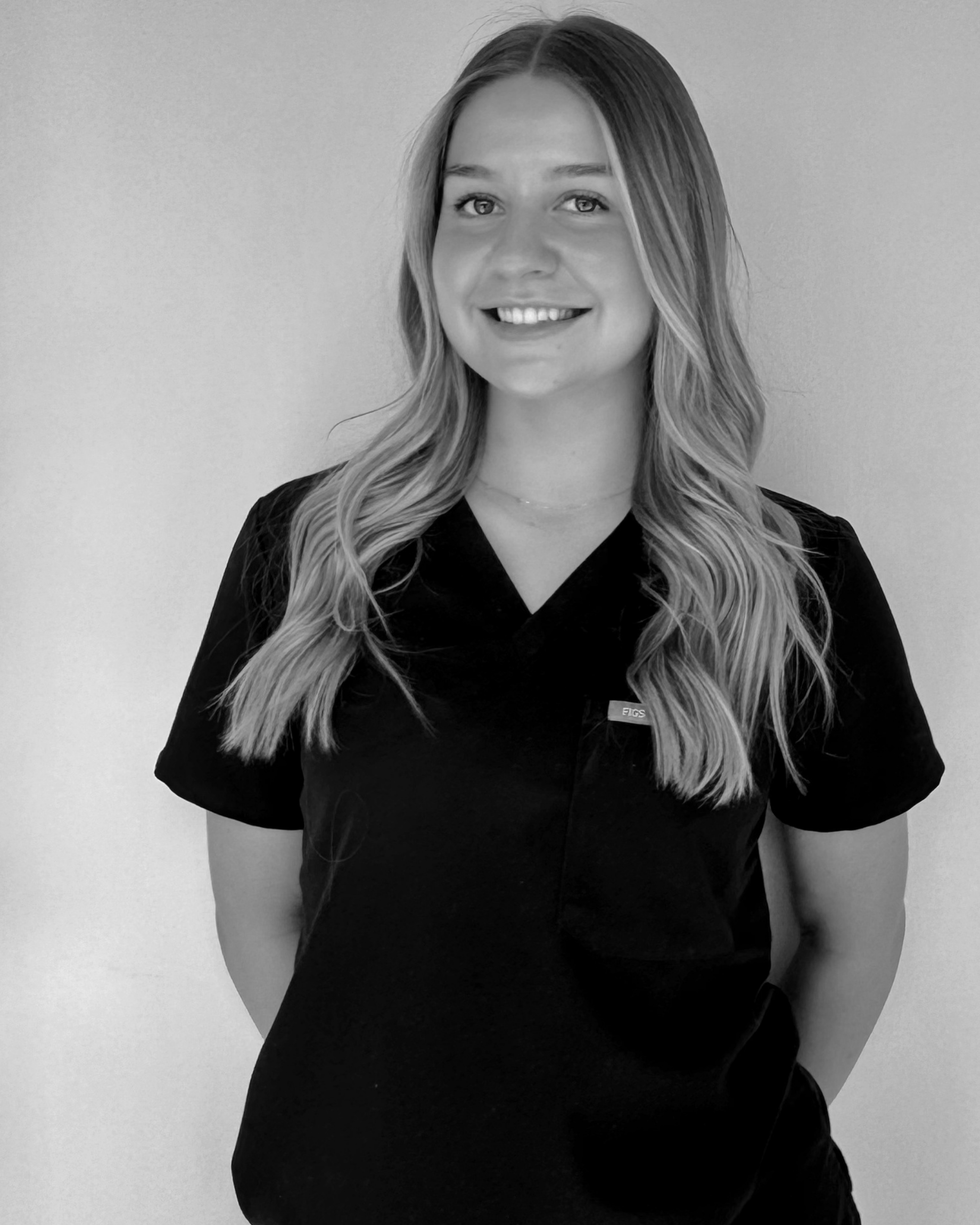 A woman with long, wavy hair smiling, wearing a dark uniform top, standing against a plain wall.