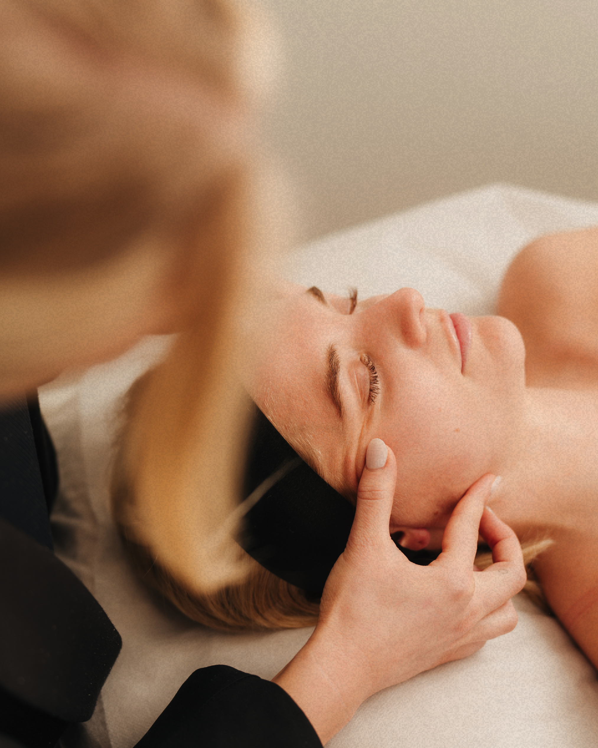 A woman lying down with her eyes closed while a person provides facial massage or therapy.