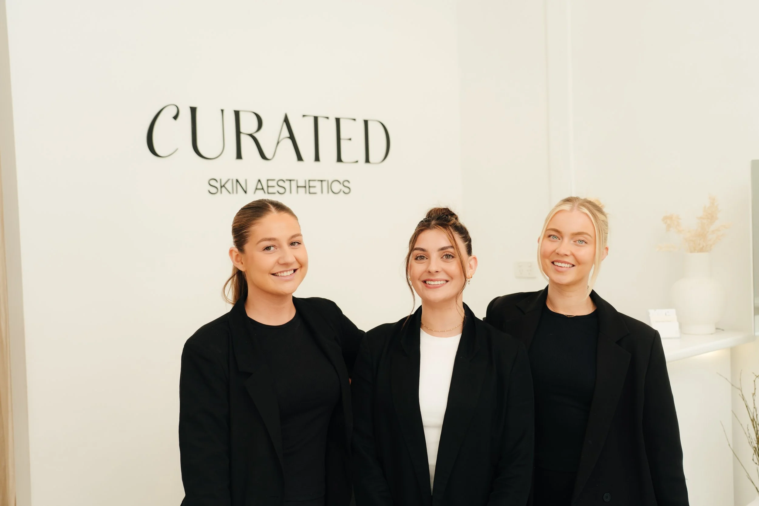 Three women standing and smiling in front of a wall with the sign 'Curated Skin Aesthetics' in a modern, minimalist interior.