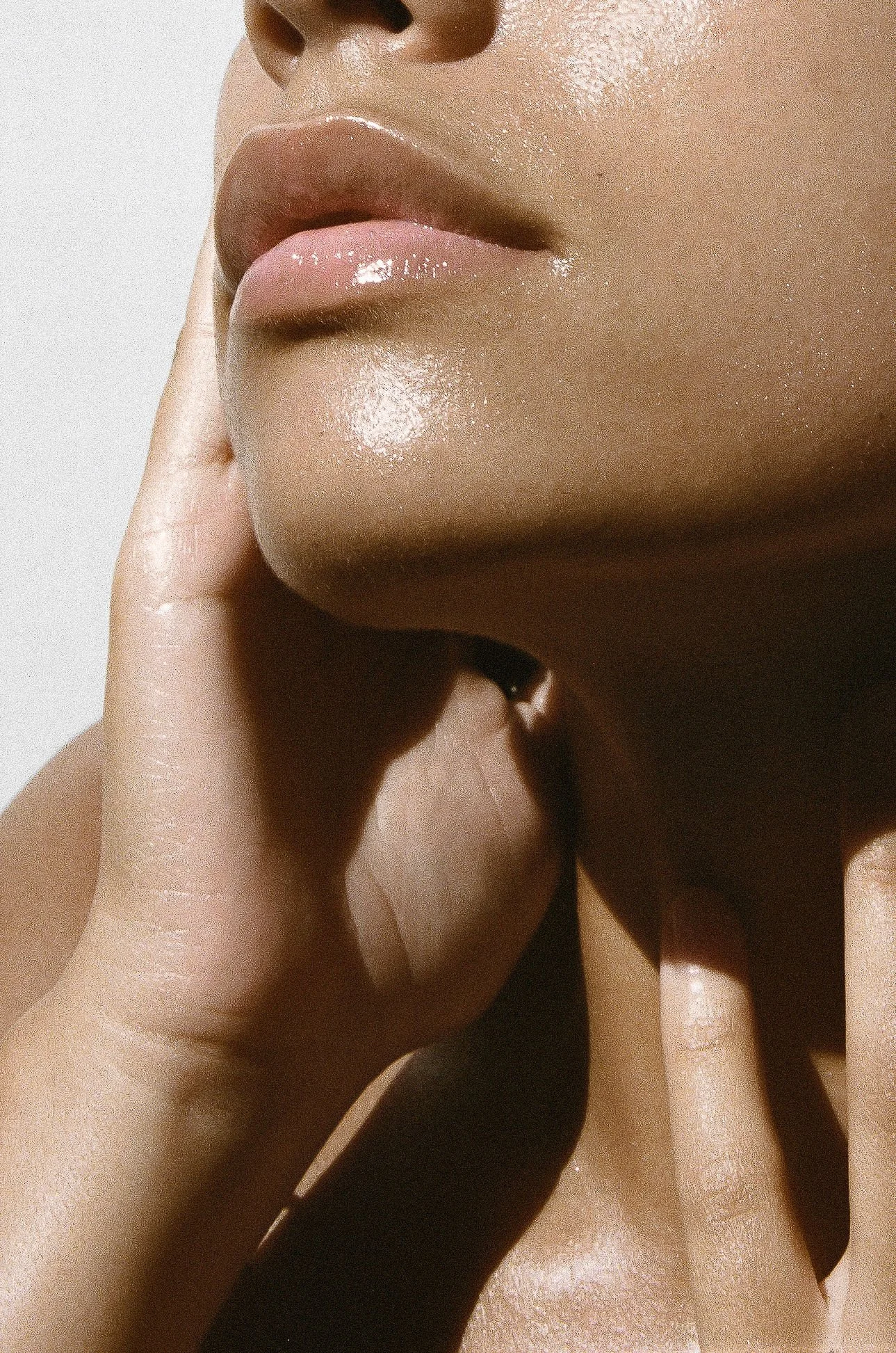 Close-up of a person's lips, chin, and neck, with a hand gently touching the face. The person has smooth, tan skin with a glossy finish on the lips and chin.