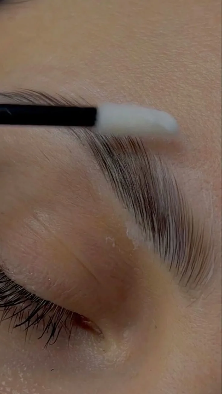 Close-up of a person applying eyelash extension adhesive to their upper eyelashes with a small applicator tool.