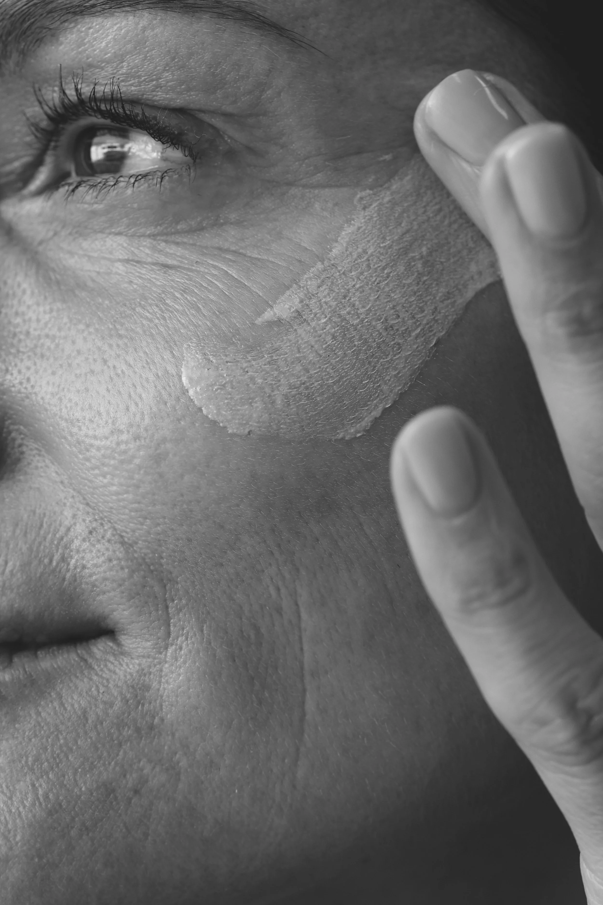 Black and white close-up of a person peeling off a facial mask from their cheek with their fingers.