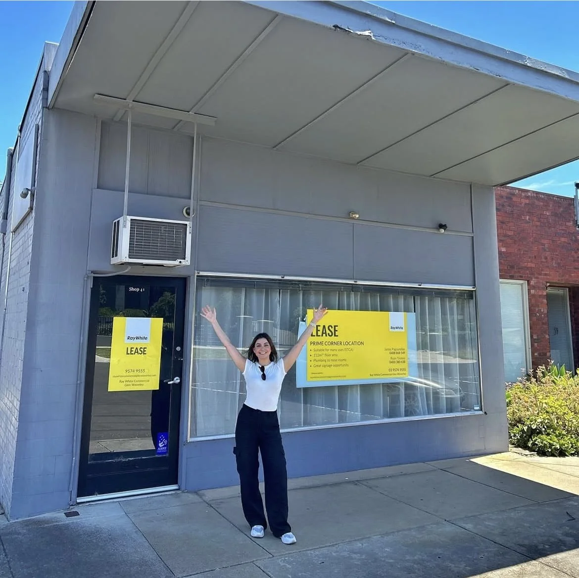 A woman standing outside a commercial building with a bright yellow lease sign in the window, smiling and raising her arms, under a clear blue sky.