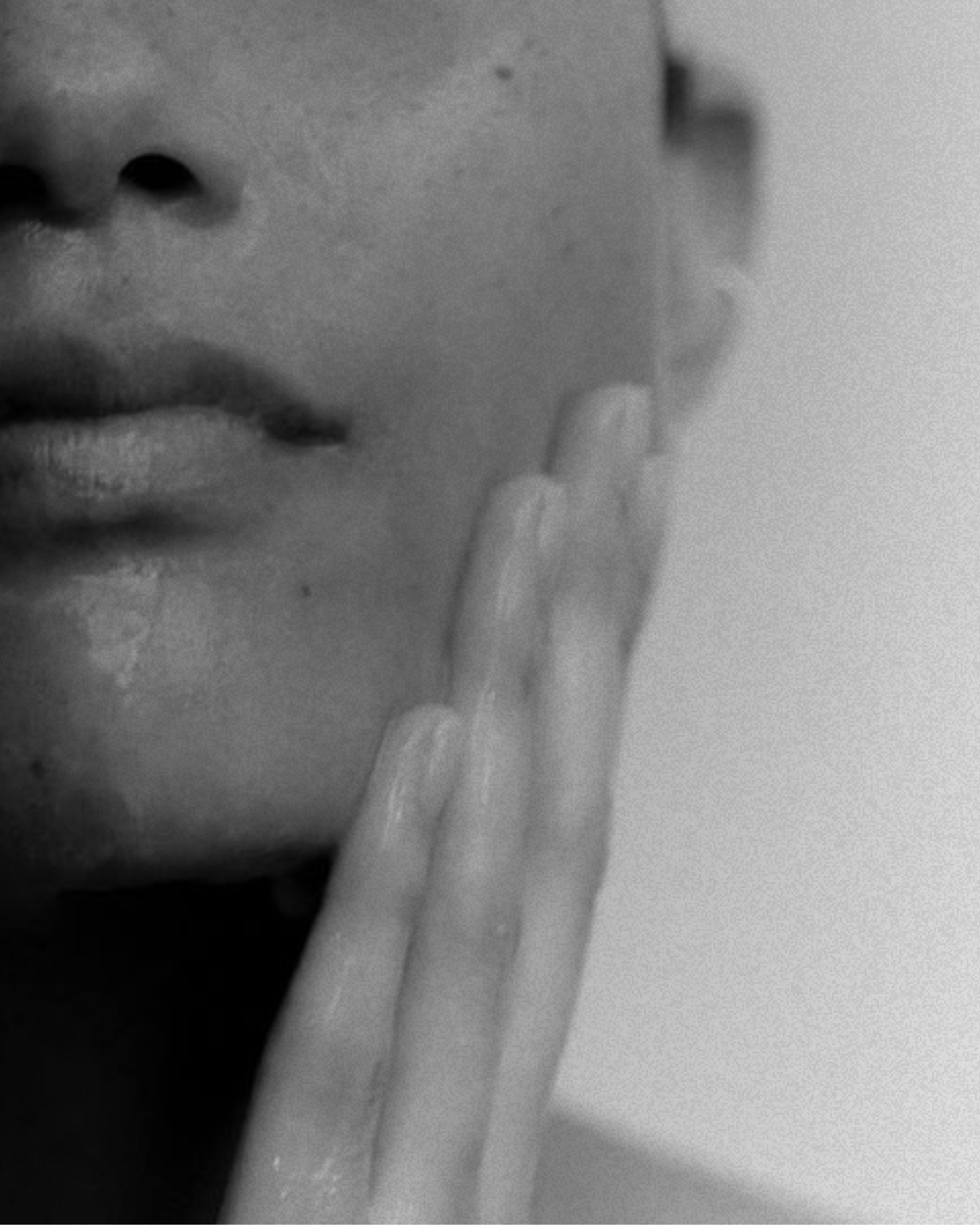 Close-up black and white photograph of person's face with fingers pressed against cheek, focusing on lips and hand.