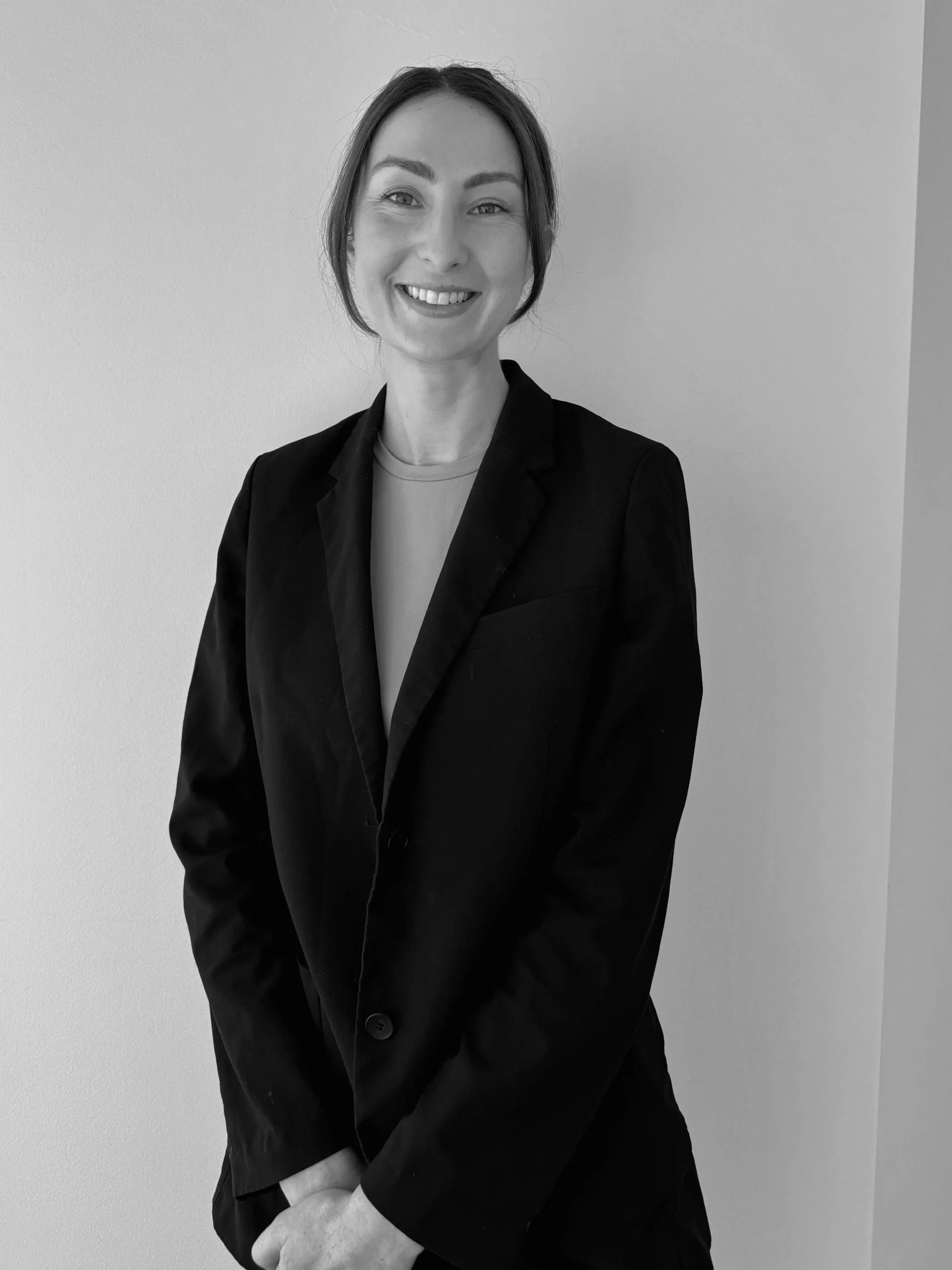 A black and white photo of a woman smiling, wearing a blazer over a blouse, standing against a plain wall.