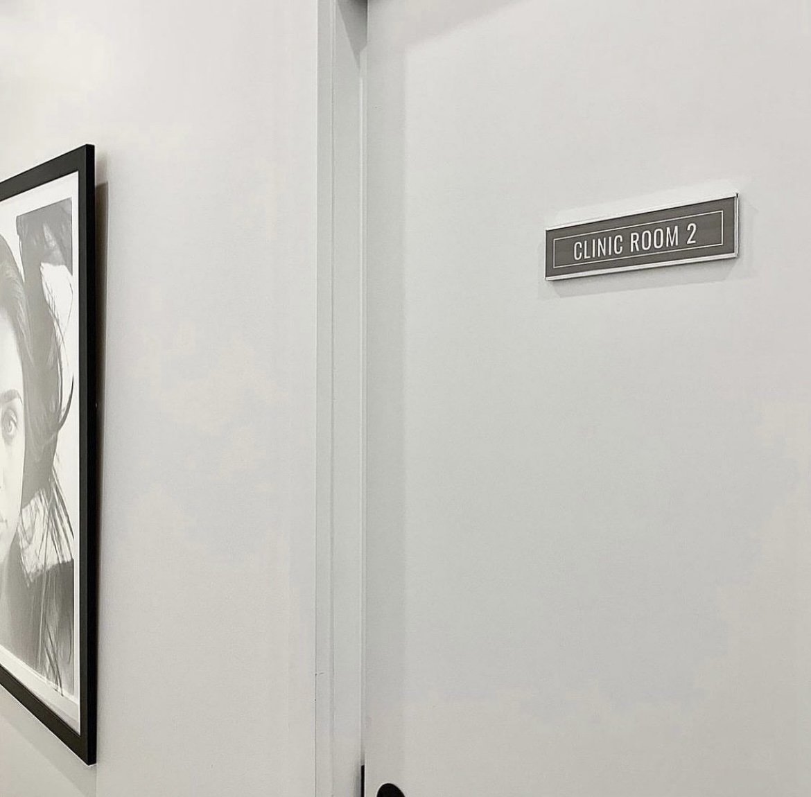 A hallway with a sign reading 'Clinic Room 2' and a framed black-and-white portrait on the wall.