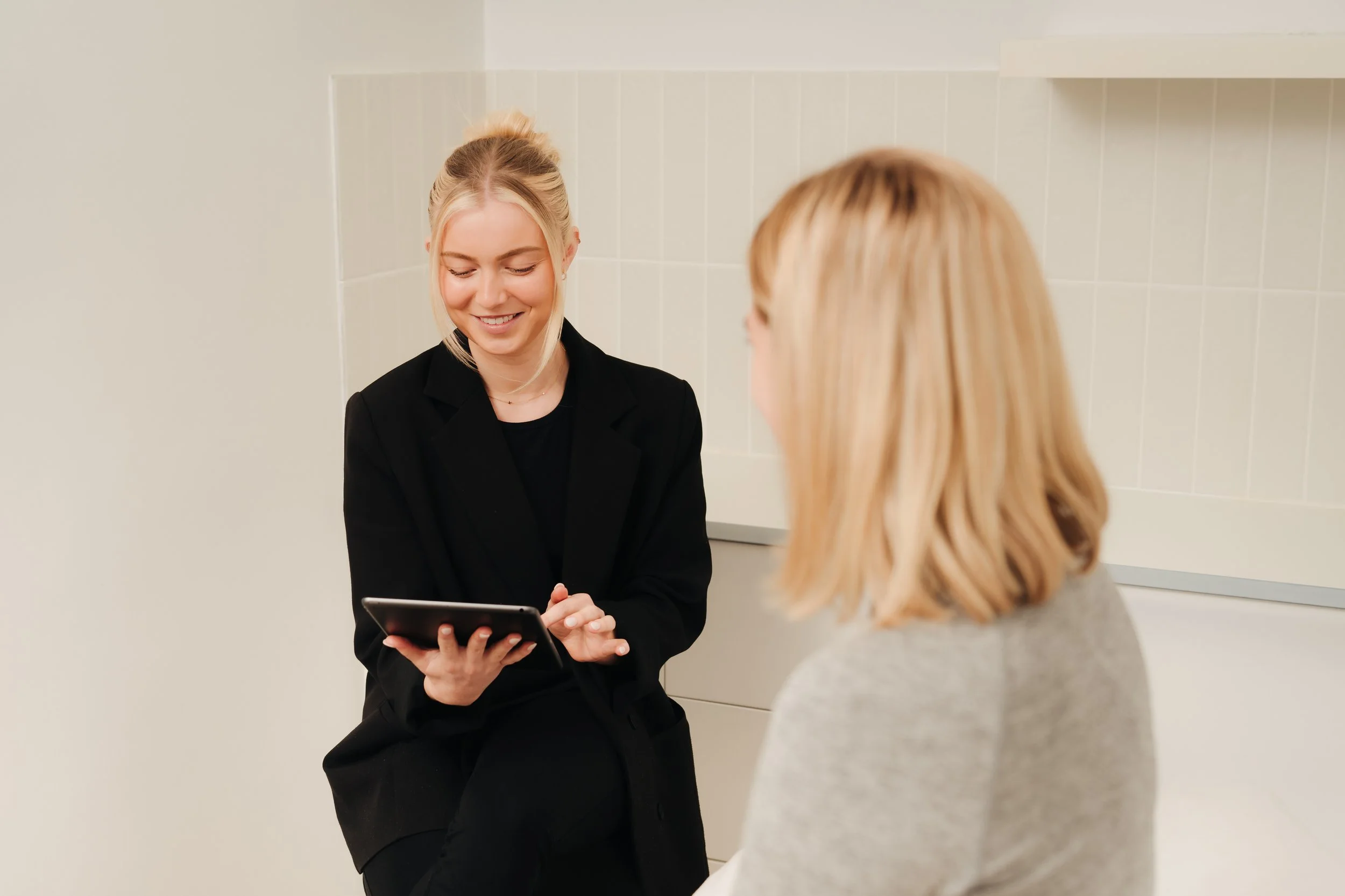 Two women sitting and engaging in a conversation, one woman is smiling while looking at a tablet device in her hands. The background is plain with white tiled walls.