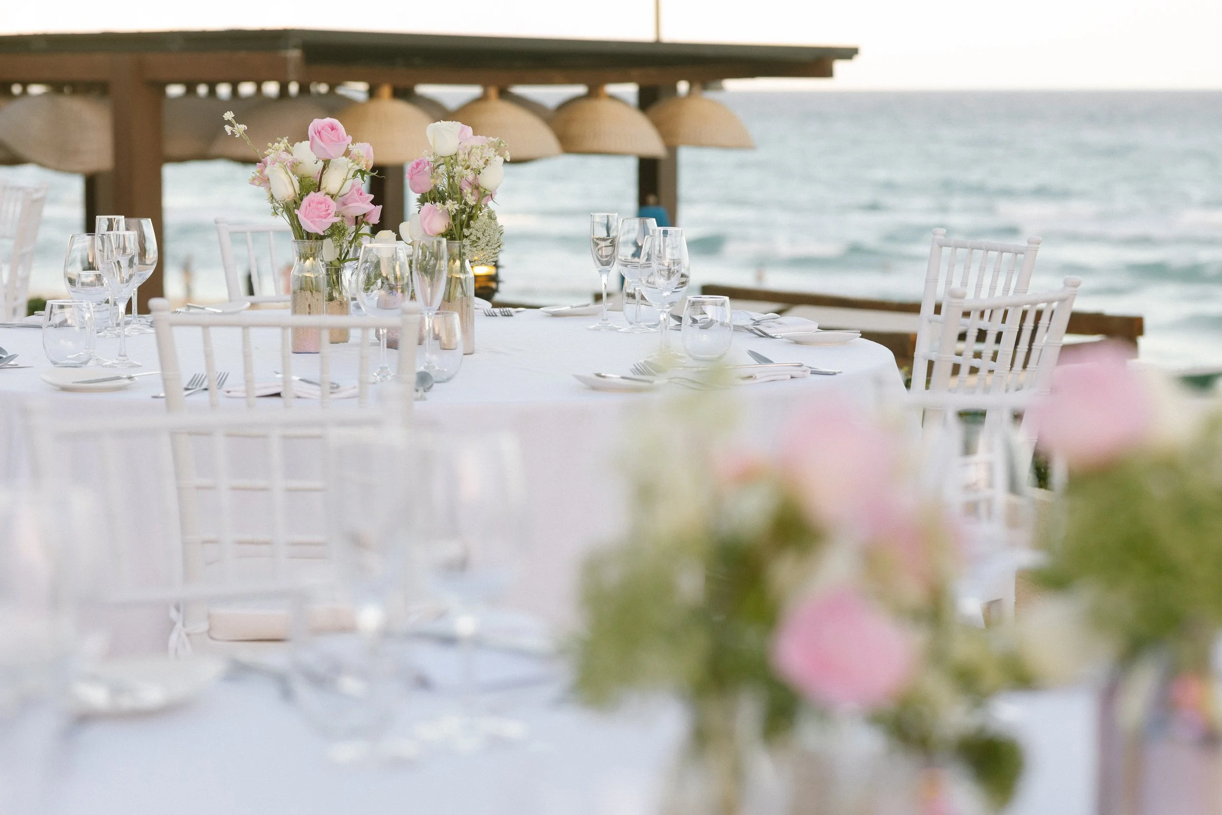 Dining table set with glassware and flower centerpieces on a deck overlooking the ocean.
