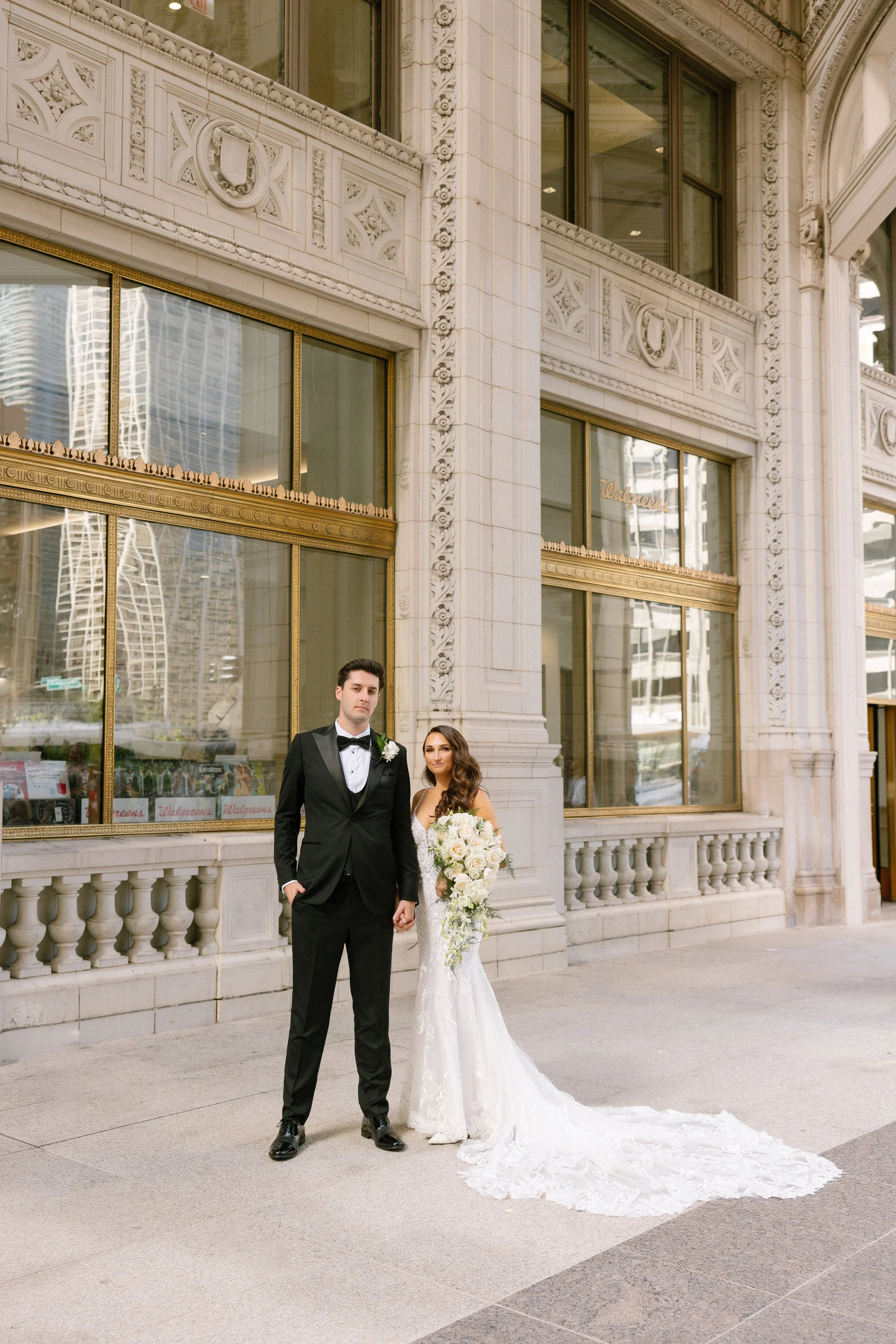 Bride and groom standing outside a grand, ornate building in wedding attire, holding hands, with the bride holding a large bouquet of white flowers and wearing a long lace train.