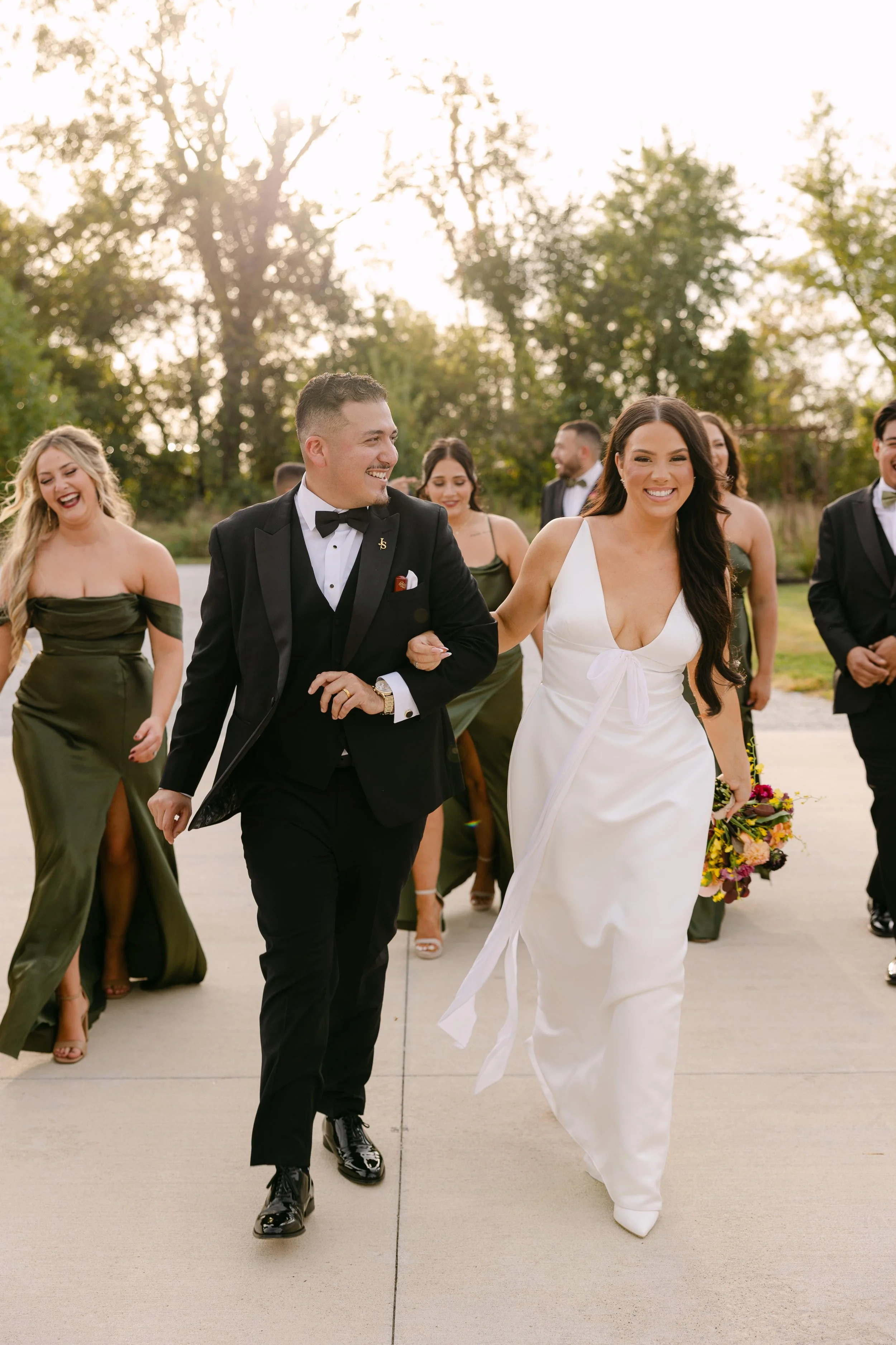 A bride and groom walking arm in arm with friends during a wedding celebration outdoors in the late afternoon. The bride is wearing a white gown, and the groom is in a black tuxedo. Guests are dressed in formal attire.