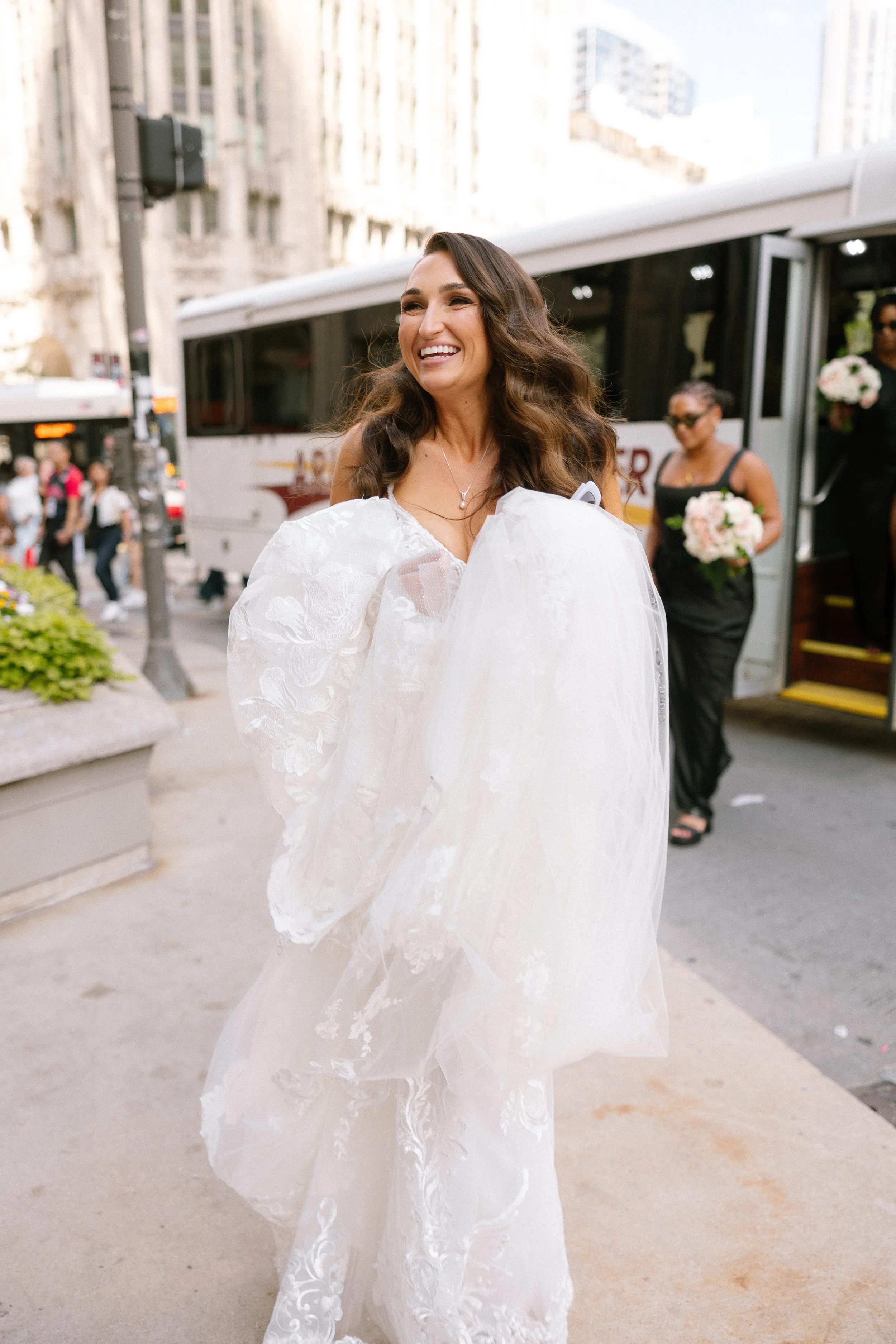 A woman in a white wedding dress smiling as she walks on a city sidewalk, with a bus and other women holding bouquets in the background.