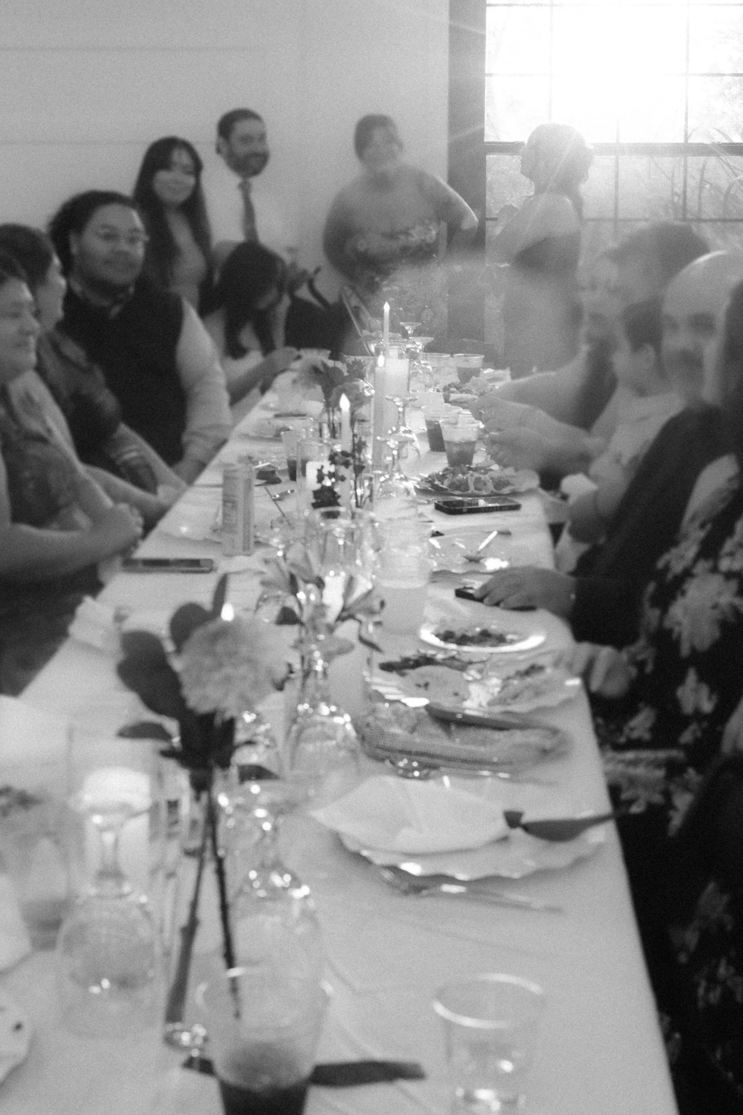 People gathered around a dining table with food, drinks, and lit candles in a bright room with large windows, during a social gathering or celebration.