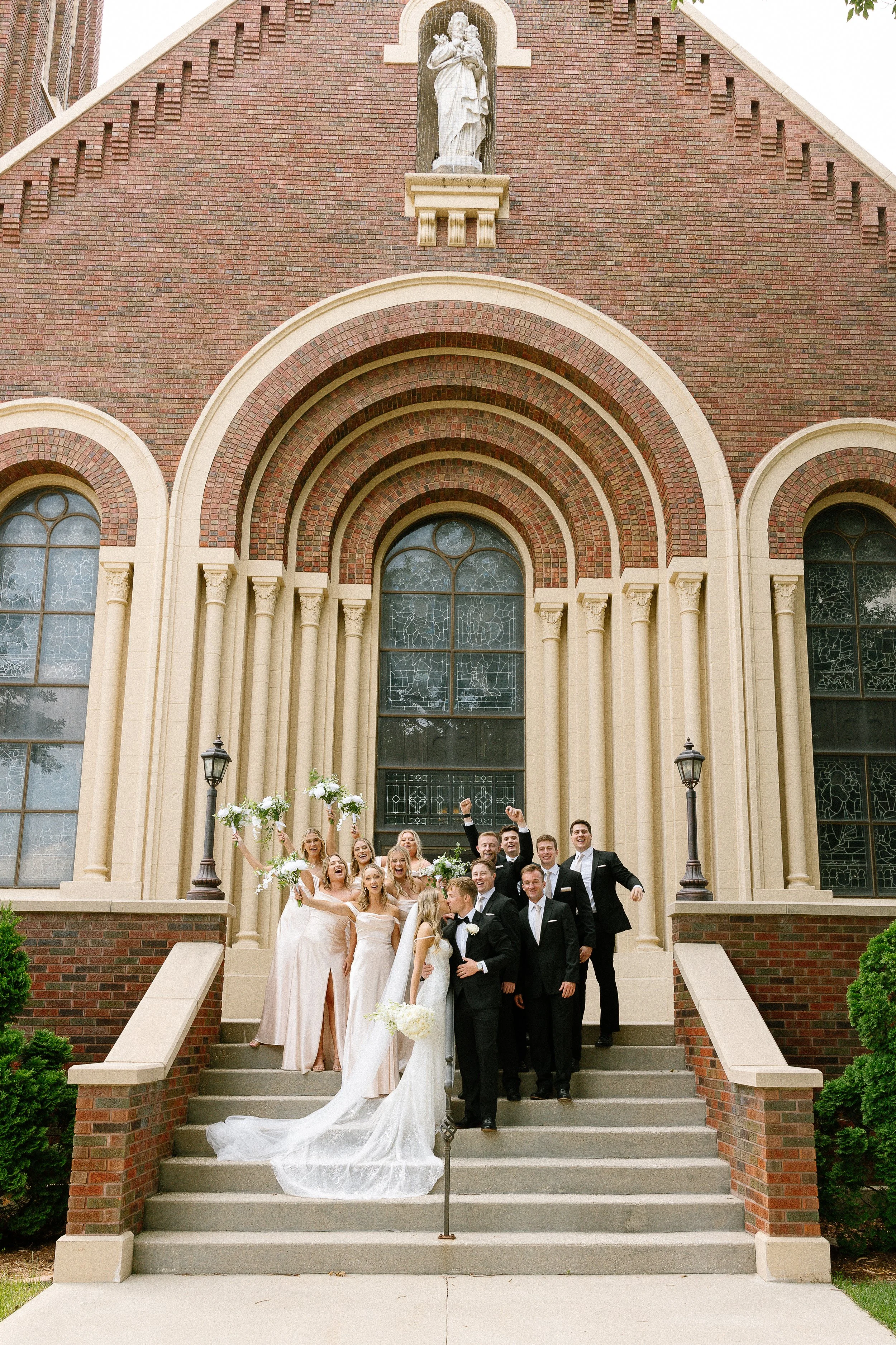 A group of wedding guests in formal attire celebrating on the steps of a brick church with arched windows and a statue of a saint above the entrance.