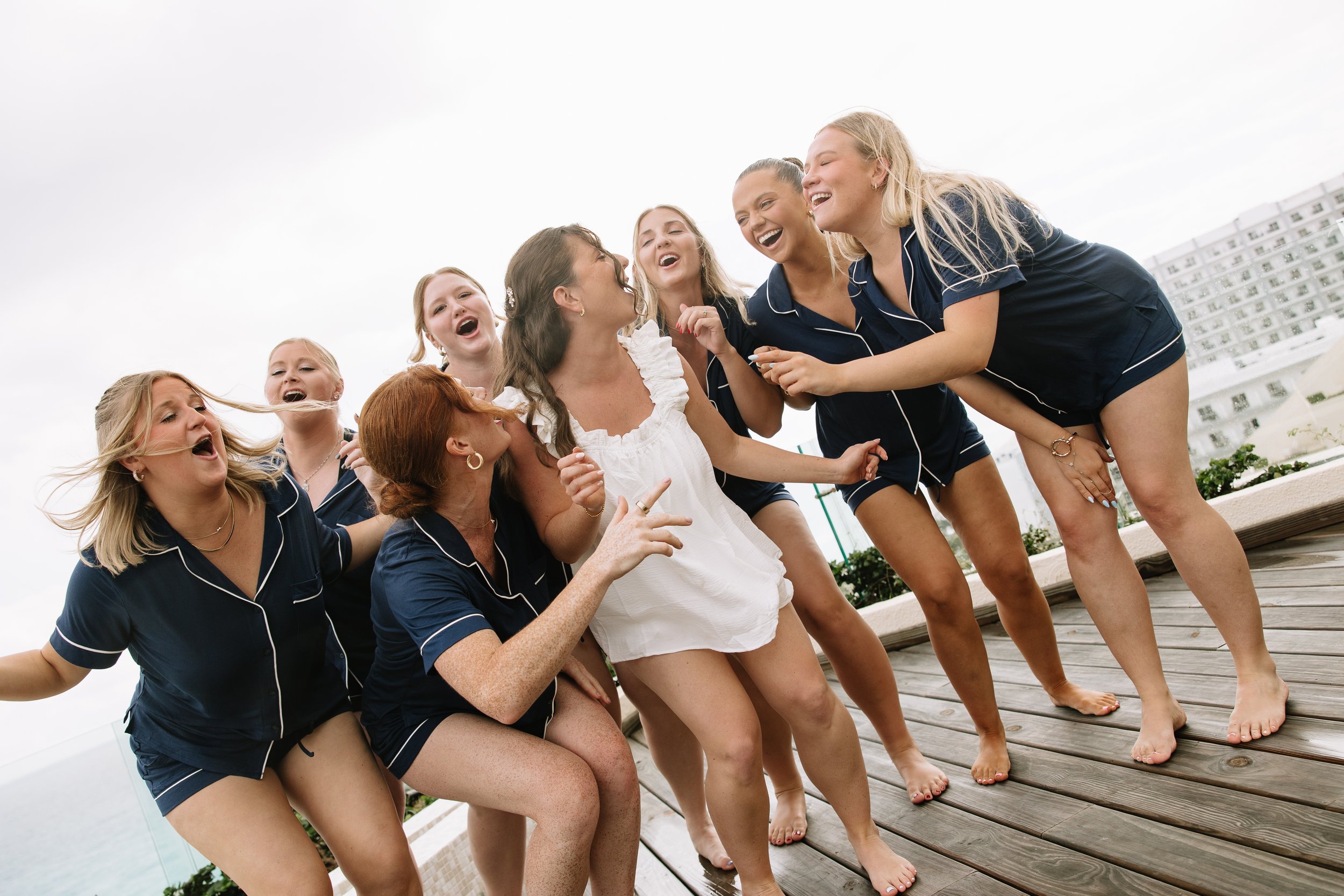 A group of women in navy pajamas laughing and playing together on a rooftop deck with an ocean view and city buildings in the background.