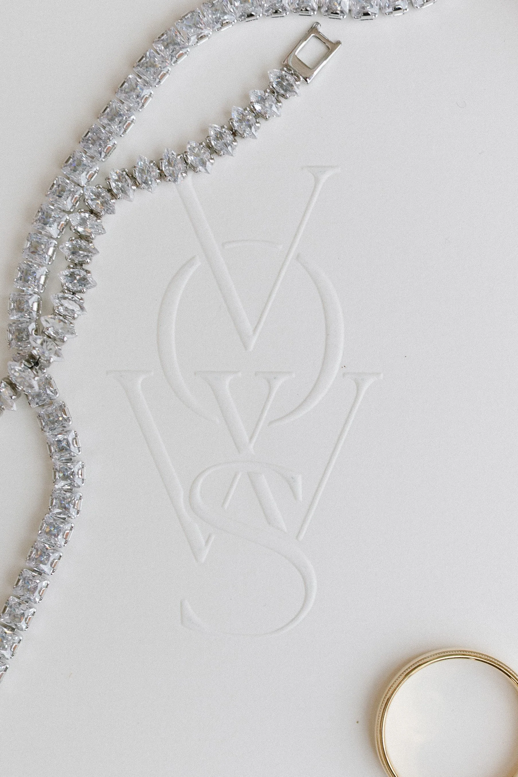 A diamond tennis necklace, a gold ring, and a white embossed card with overlapping V, W, and S letters