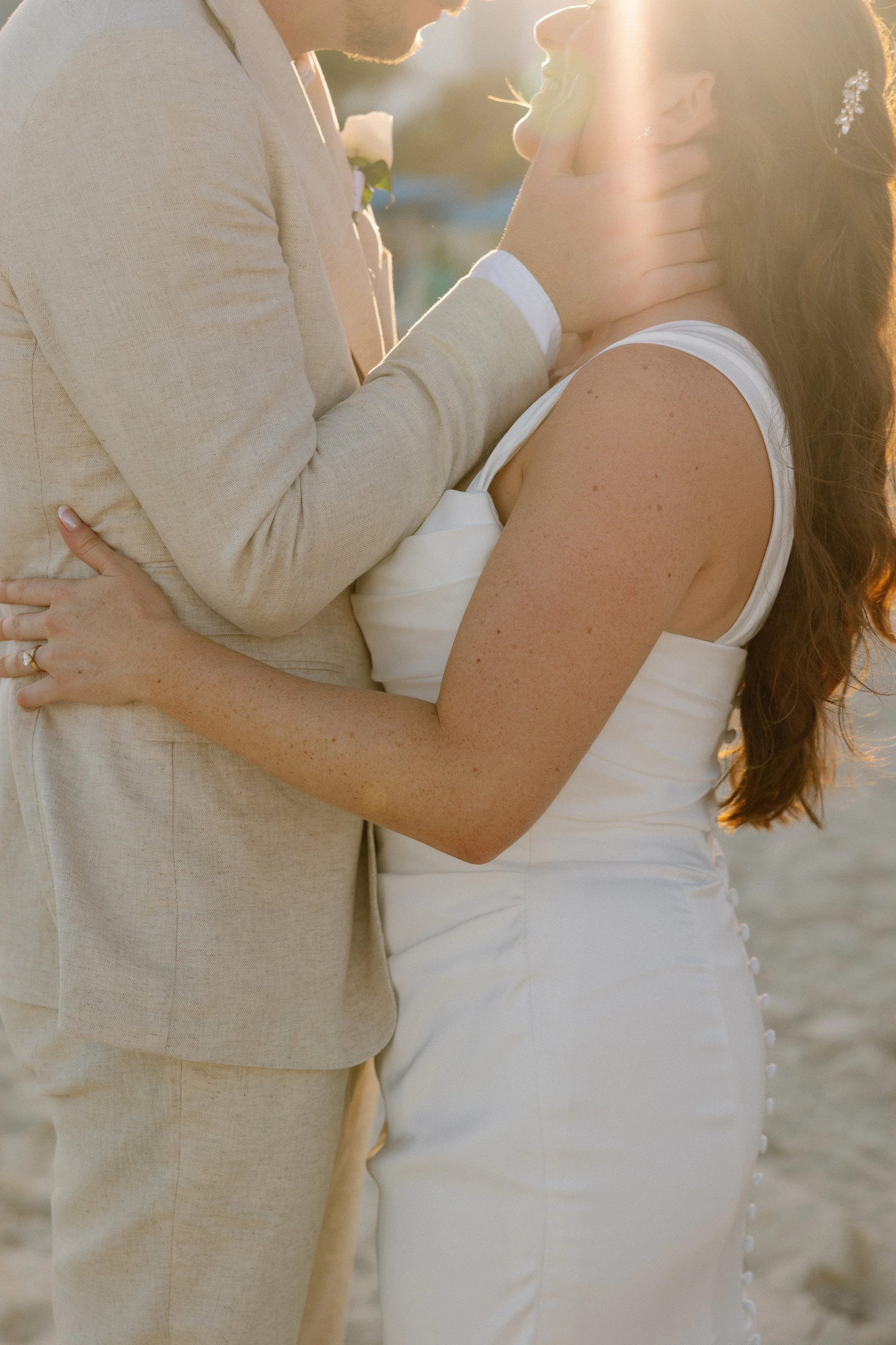 A couple in wedding attire embraces on the beach during sunset, with the groom gently holding the bride's face, both smiling softly.