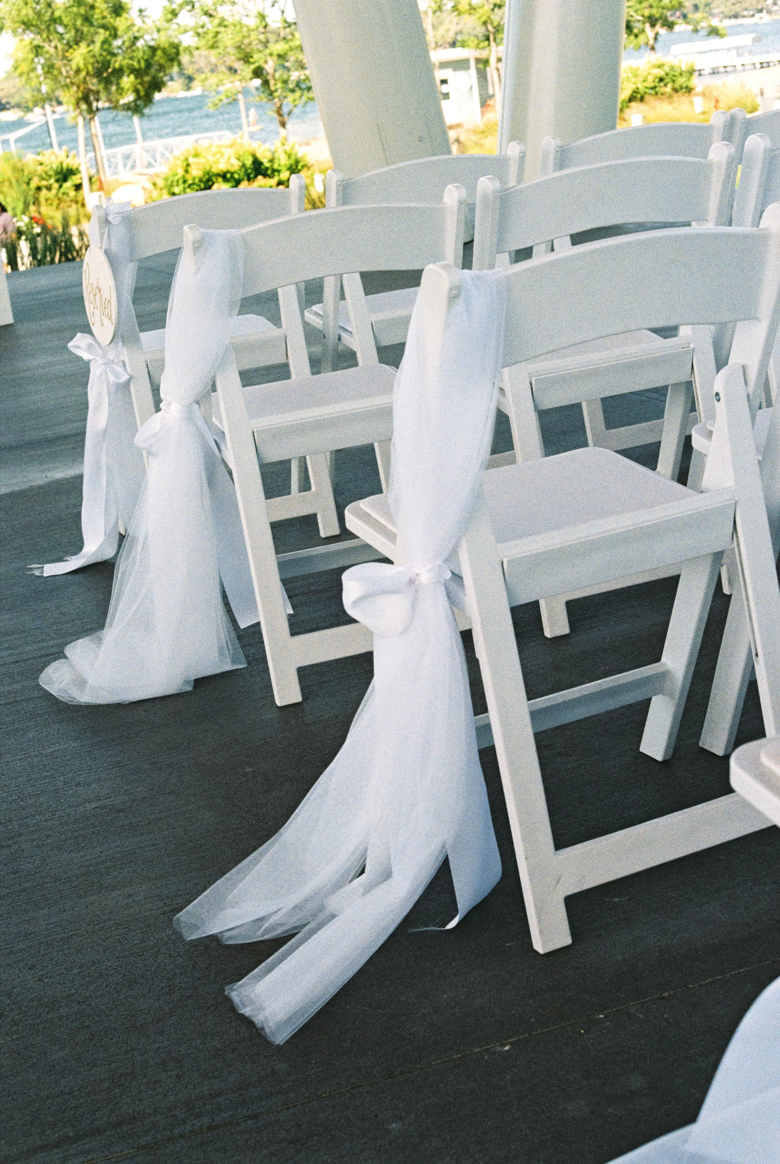 White chairs decorated with white fabric ribbons, arranged outdoors near a body of water, likely set up for a wedding ceremony.
