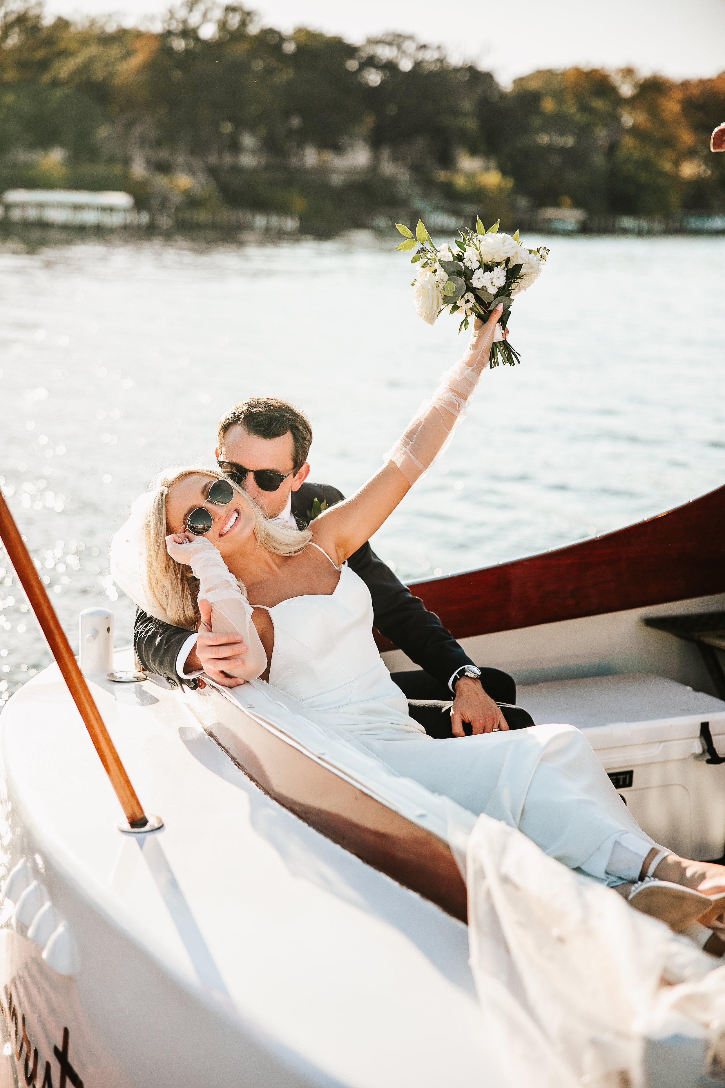 A happy bride and groom on a boat, with the bride holding a bouquet of white flowers and raising her arm, wearing sunglasses and a white wedding dress, during sunset on the water.