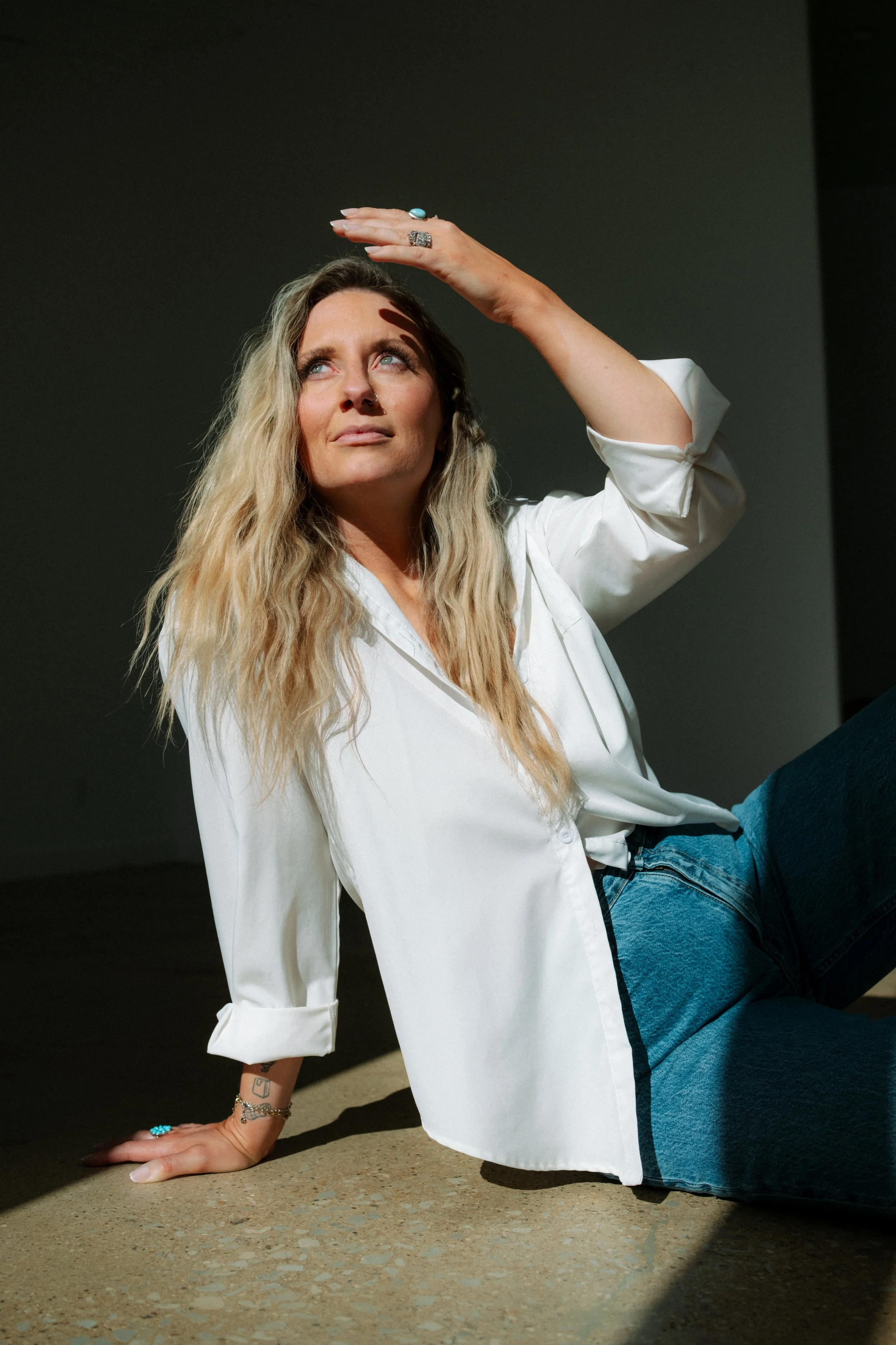 Woman with long wavy blonde hair and blue eyes, sitting on the floor with one hand supporting her and the other hand shielding her eyes from the light, wearing a white blouse and blue jeans.