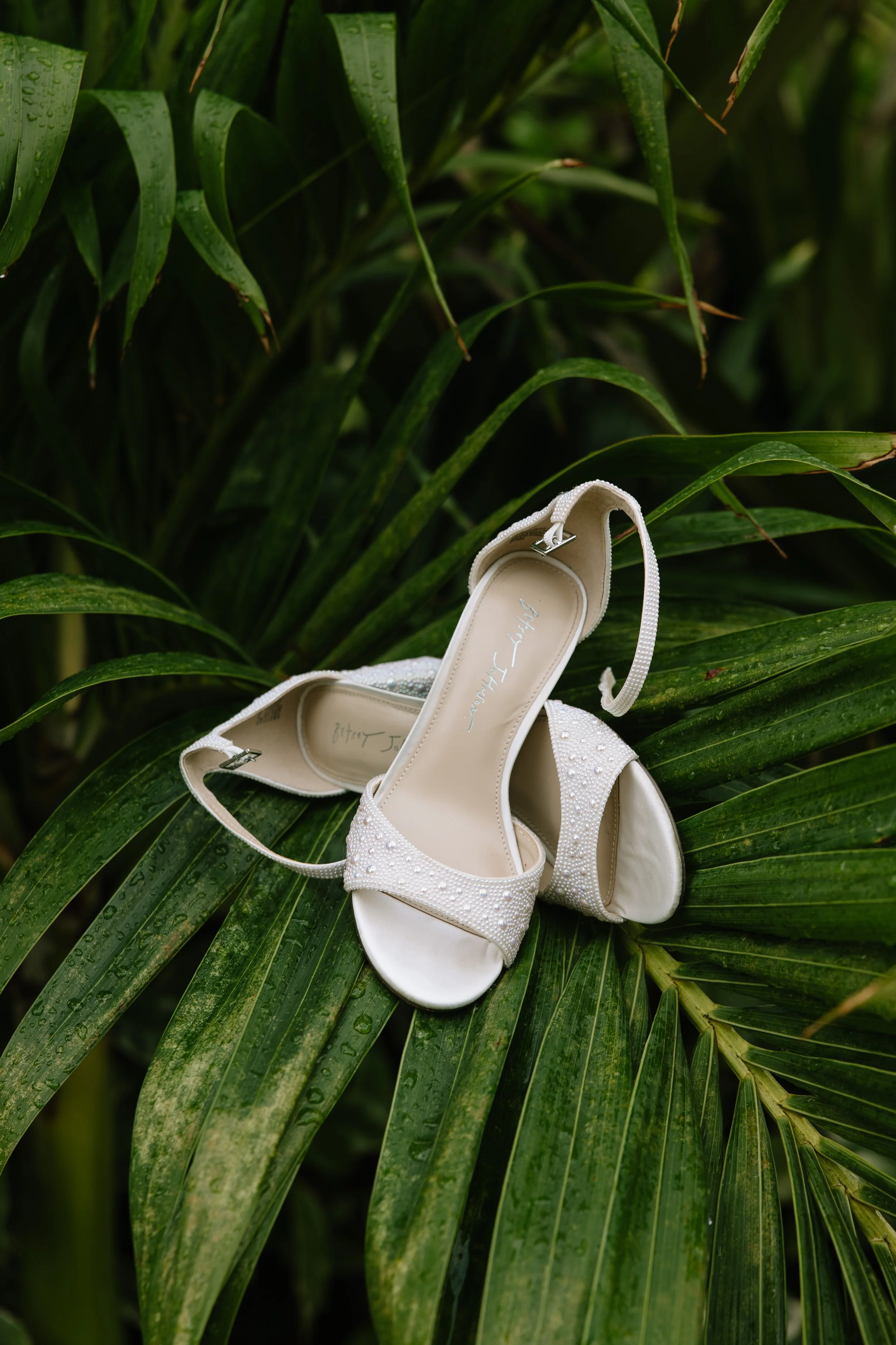 White bridal shoes with pearl embellishments resting on green tropical leaves.