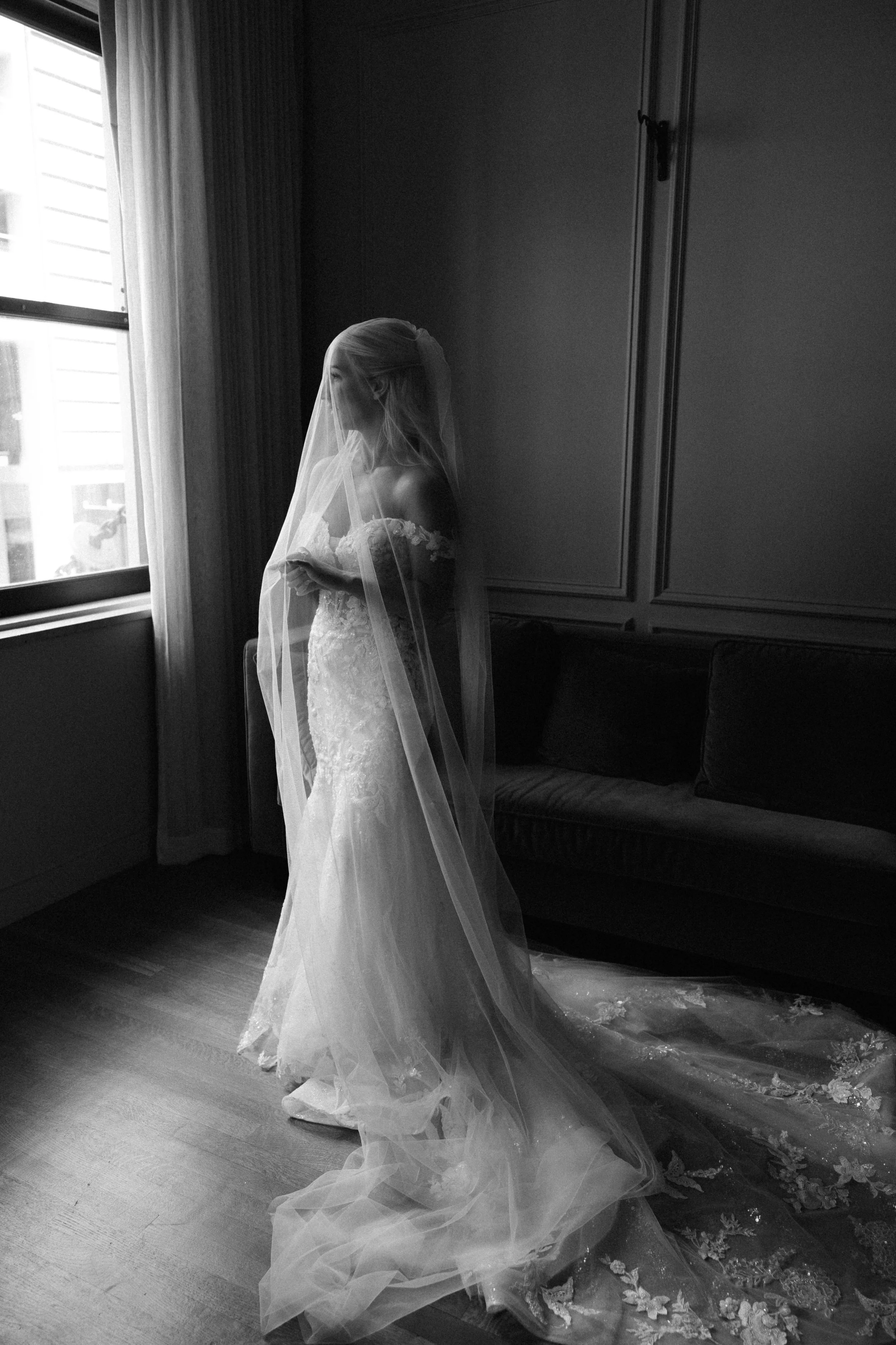 A bride in a wedding dress and veil standing by a window in a room, looking down, with a dark sofa and wall panel behind her.