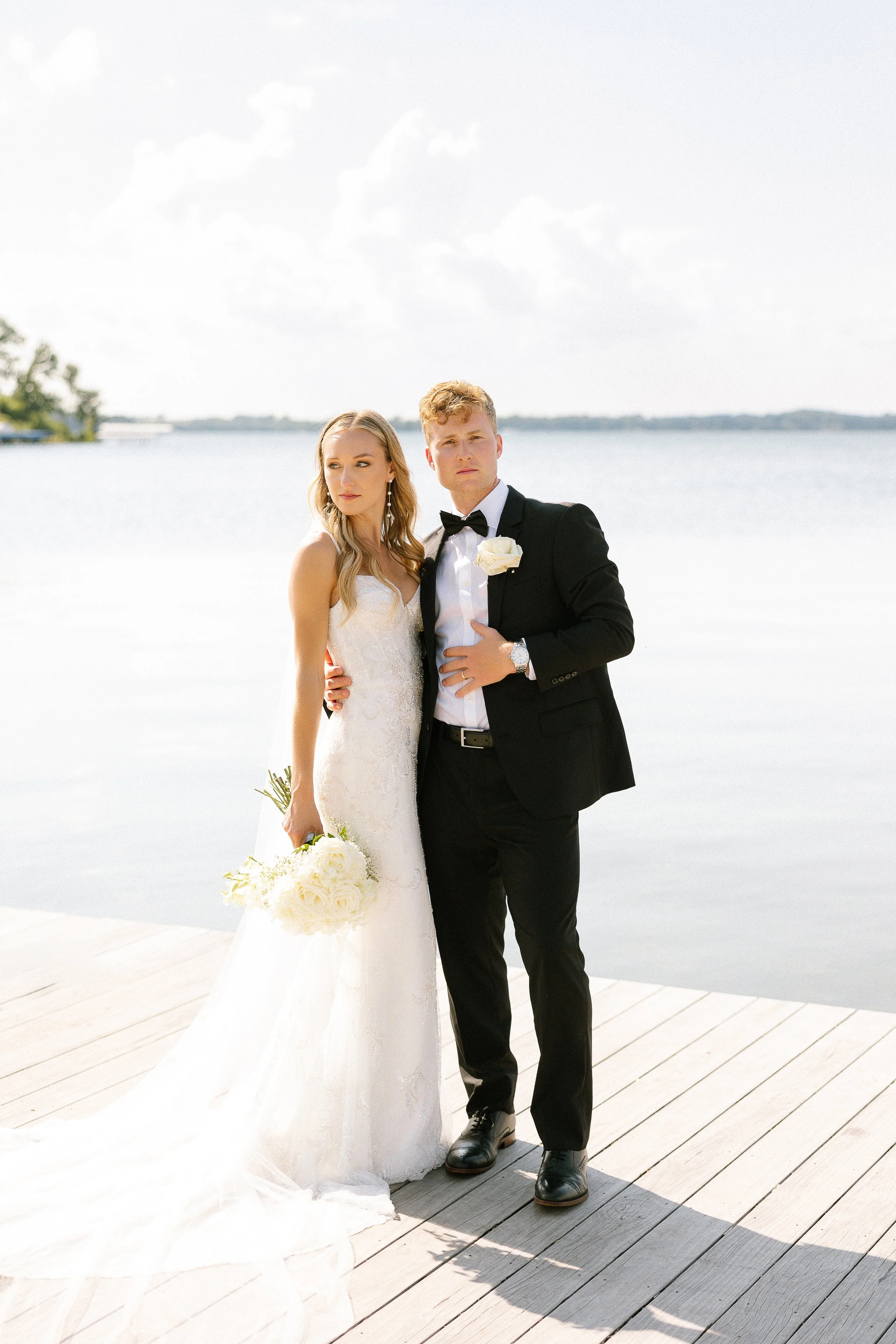 A bride and groom standing on a wooden dock by a lake, dressed in wedding attire, with the bride holding a bouquet of white roses and the groom with his hand on his chest, both looking at the camera.