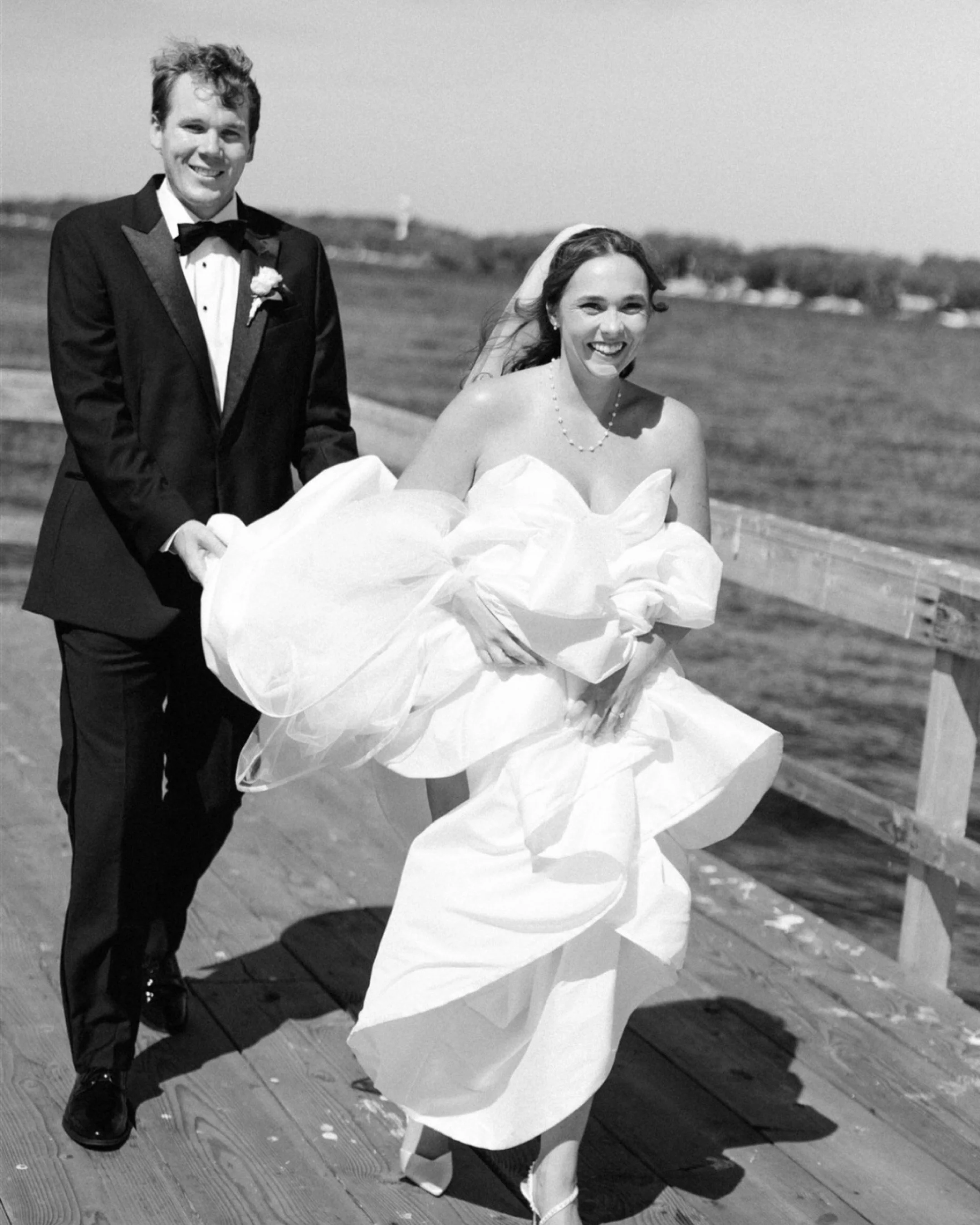 A black and white wedding photo of a smiling couple outdoors on a wooden deck with grassy landscape in the background. The groom is in a tuxedo holding the bride's hand, and the bride is in a wedding gown holding a bouquet, with a veil and jewelry.