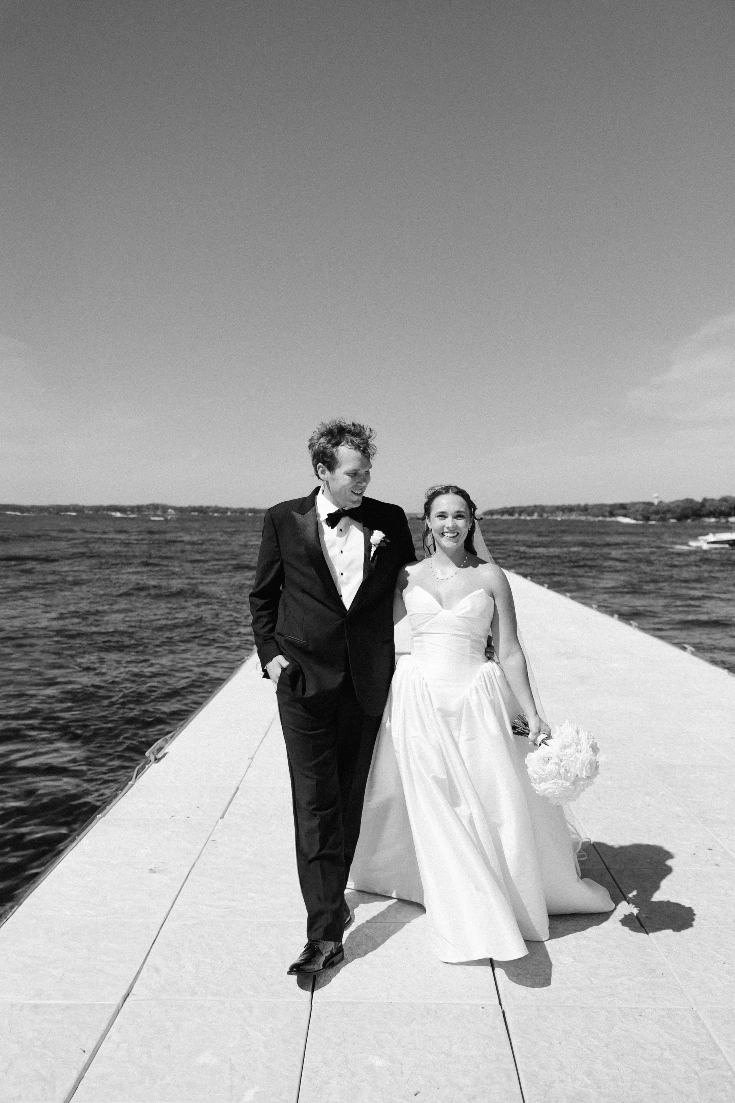 A bride and groom walking on a dock by the water, smiling, with the bride holding a bouquet, in black and white.