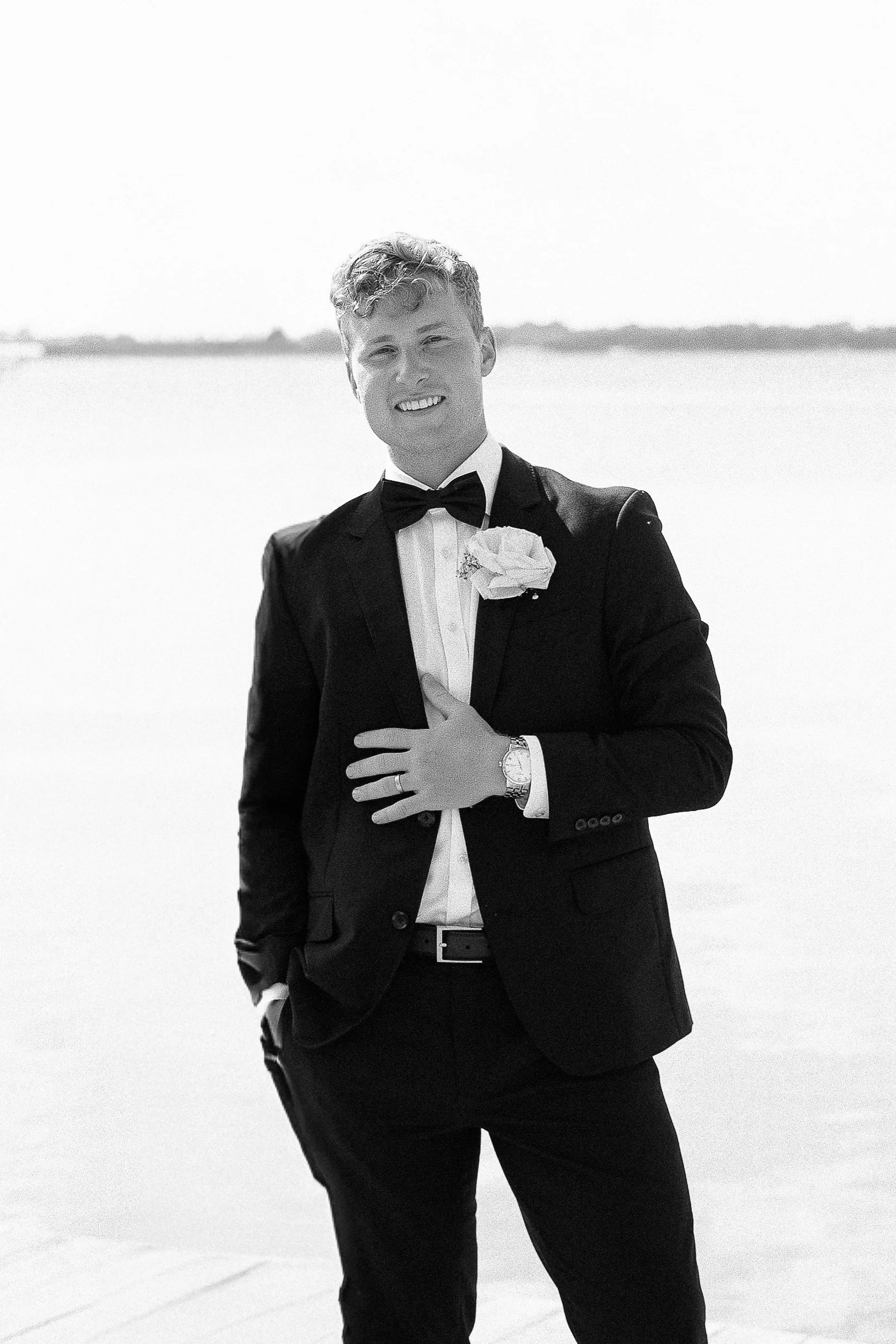 A young man dressed in a tuxedo with a bow tie and boutonnière, standing outdoors near a body of water, smiling at the camera.