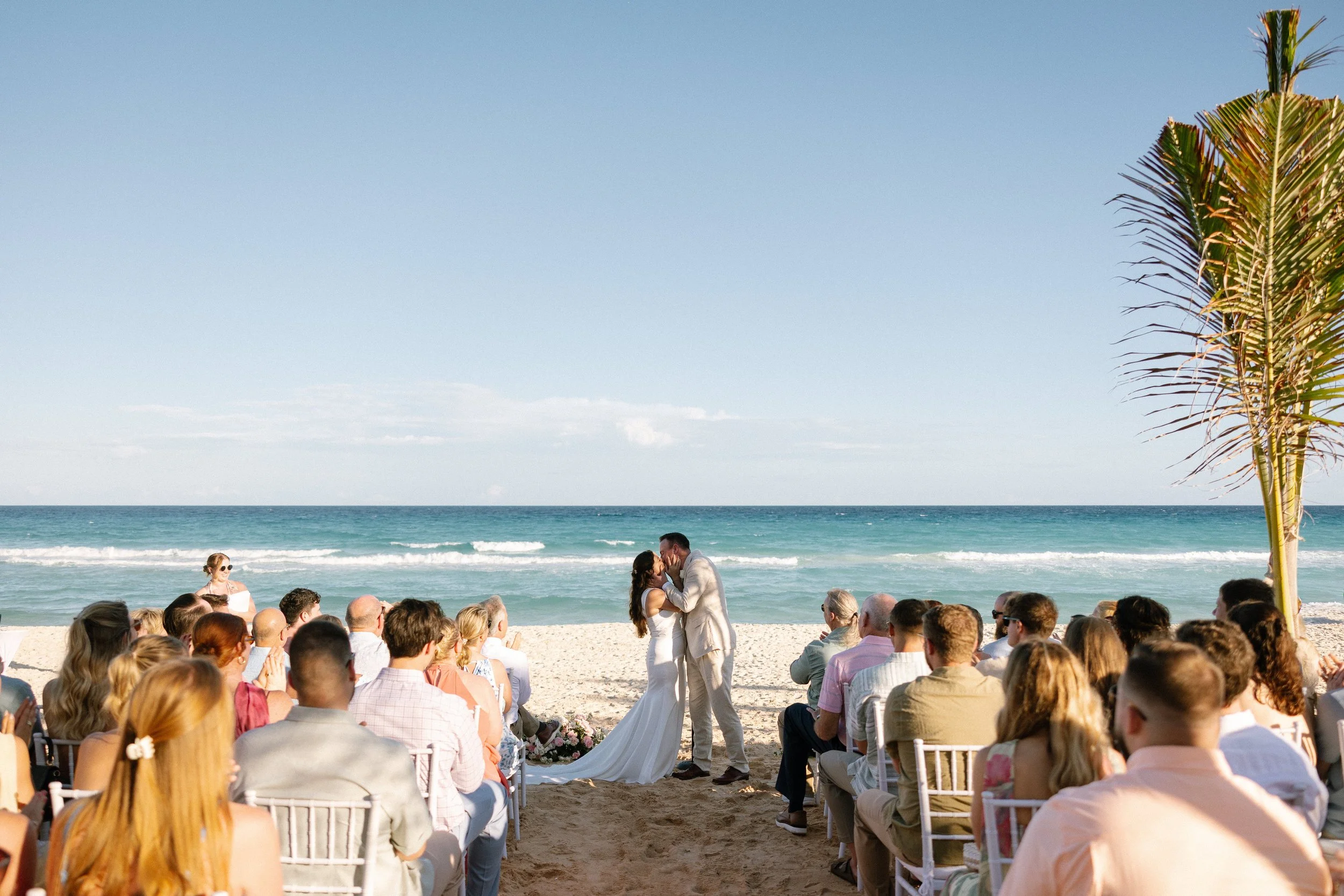 A wedding ceremony on the beach with a couple kissing in front of seated guests, ocean waves in the background, and a palm tree on the right.