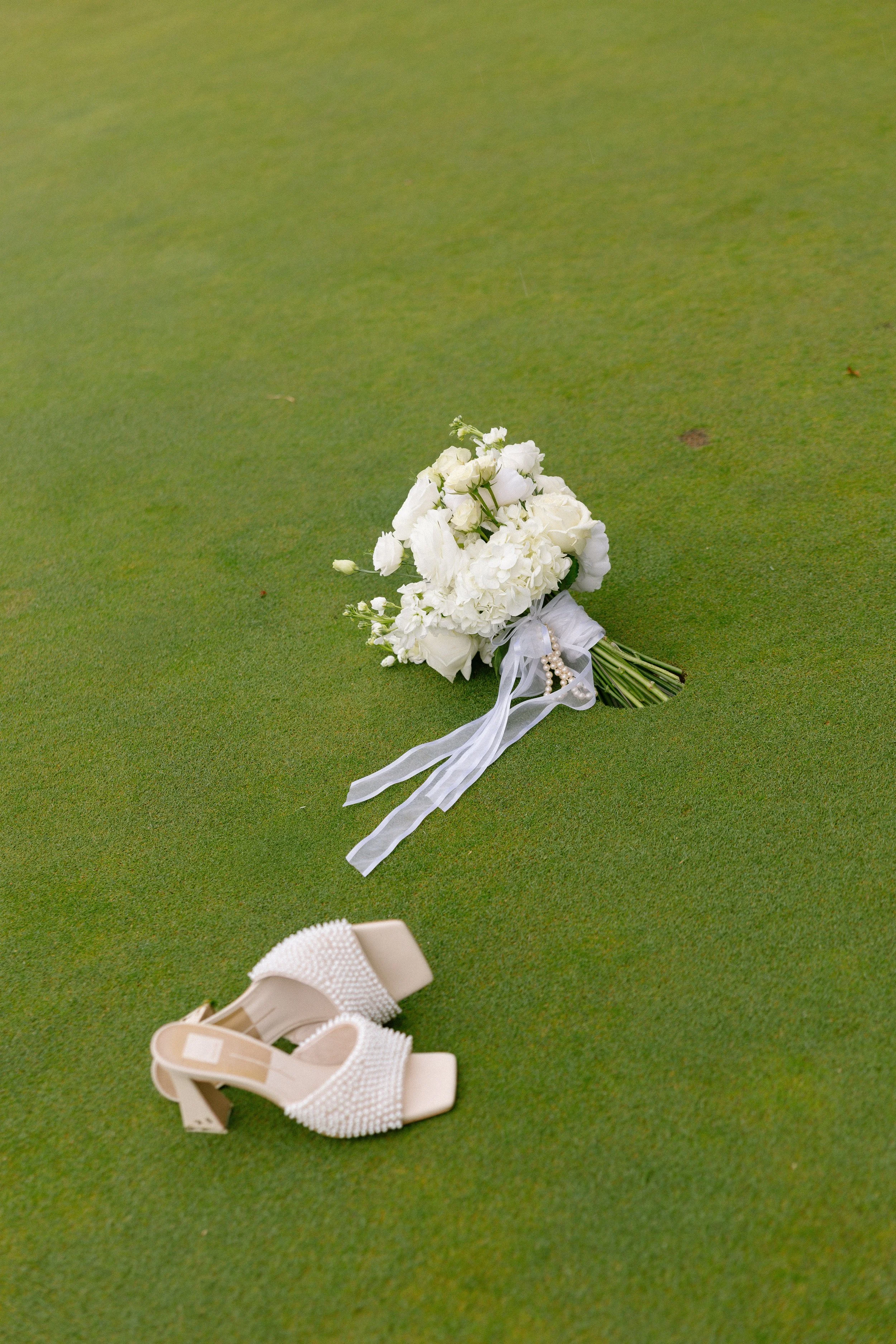 A white bridal bouquet with ribbons and a pair of white embellished high-heeled shoes on a green grass field.