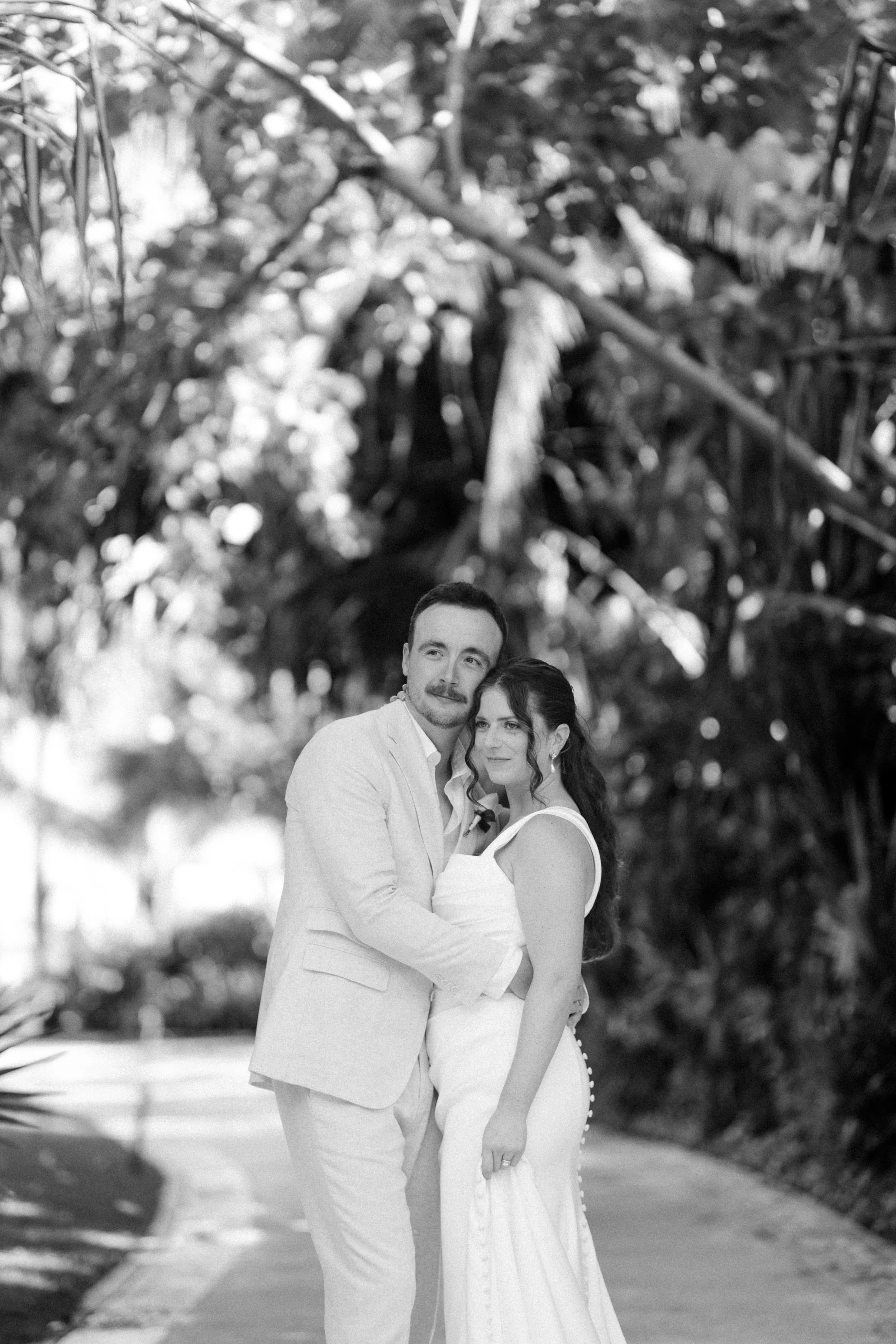 A black-and-white photo of a couple standing closely together outdoors, with trees and foliage in the background. The man is wearing a light-colored suit jacket and shirt, and the woman is in a white dress with button details on the side. They are sm