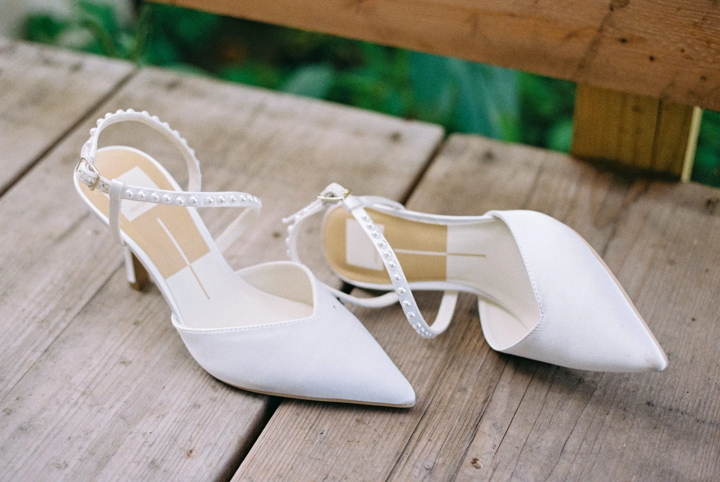 A pair of white bridal shoes with pointed toes, ankle straps with pearl embellishments, placed on a wooden surface.