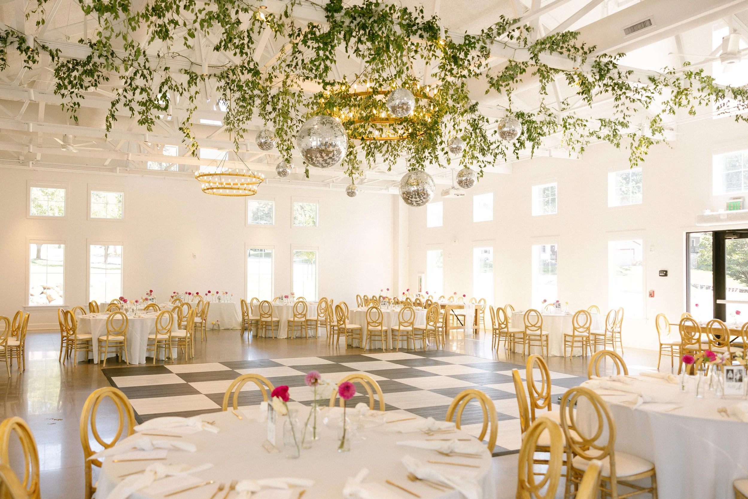 A decorated banquet hall with round tables covered in white tablecloths, adorned with pink flowers in small vases, and set with plates and napkins. The hall features high ceilings with large windows, hanging green foliage, and disco ball decorations, creating a festive atmosphere. There is a checkered dance floor in the center of the room.