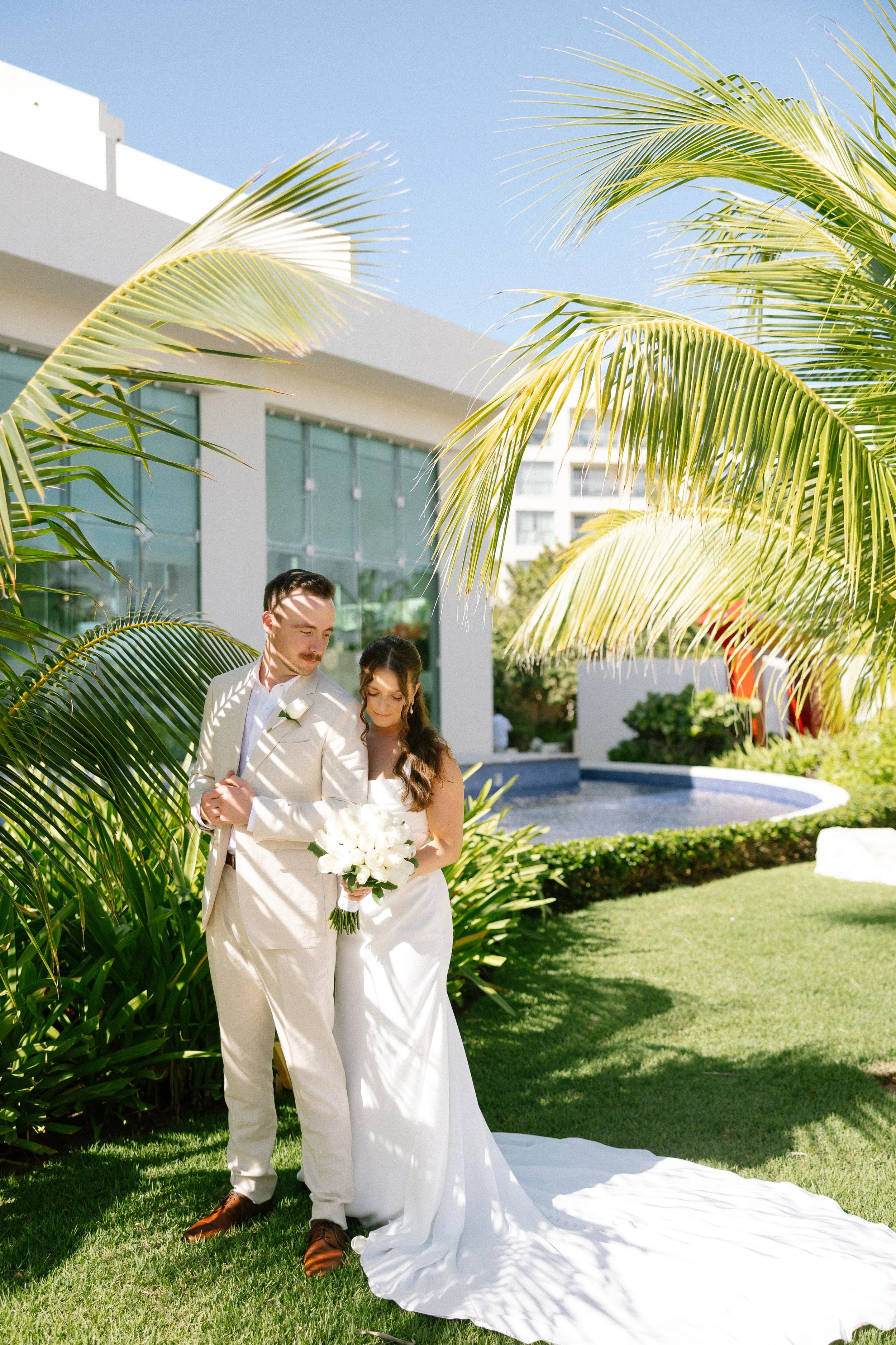 A bride and groom standing on a lush green lawn outdoors, surrounded by palm trees, with a modern building and a small fountain in the background. The bride is holding a bouquet of white flowers, and they are dressed in white wedding attire.
