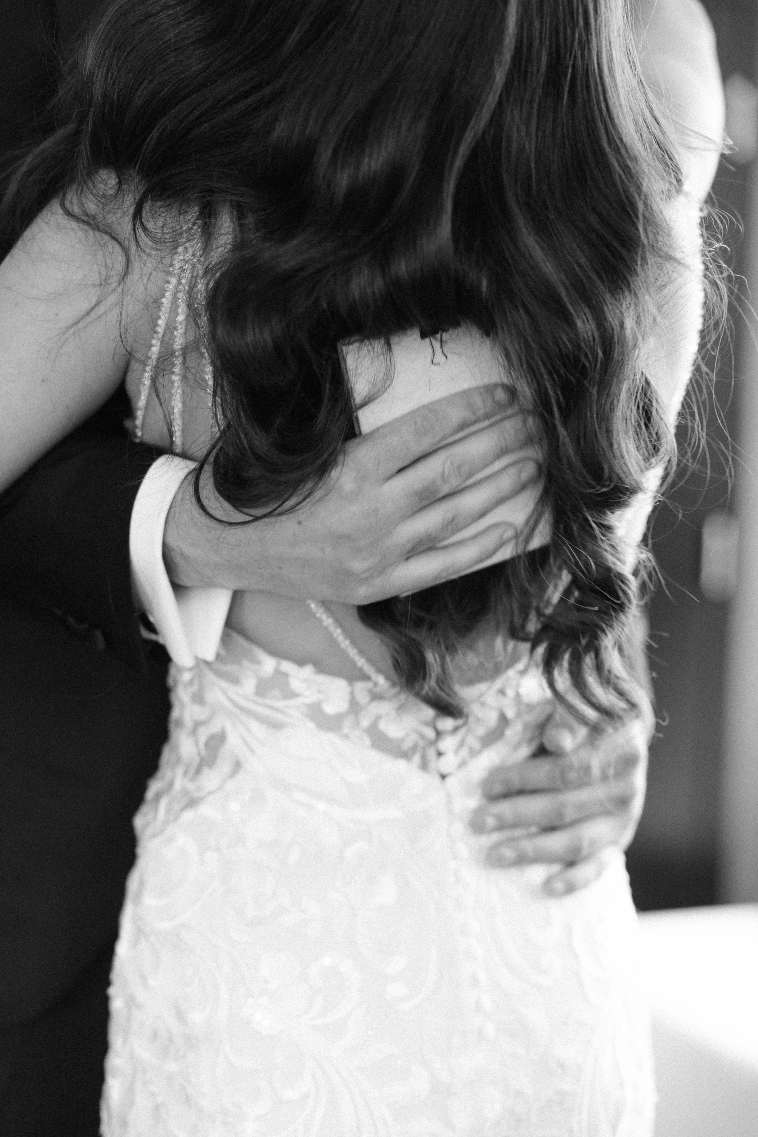 A woman in a wedding dress embracing and crying with a man in a tuxedo, her head resting on his shoulder.